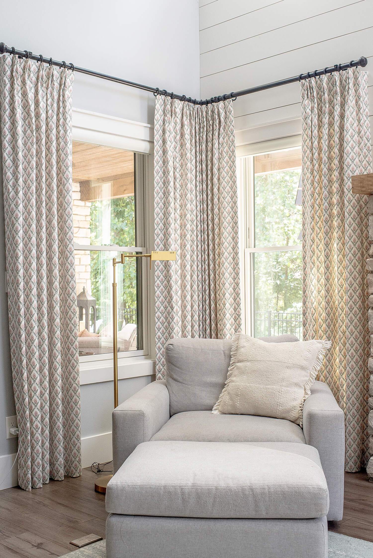 Light-filled living room with patterned drapes, gray armchair, floor lamp, and large windows for natural brightness.