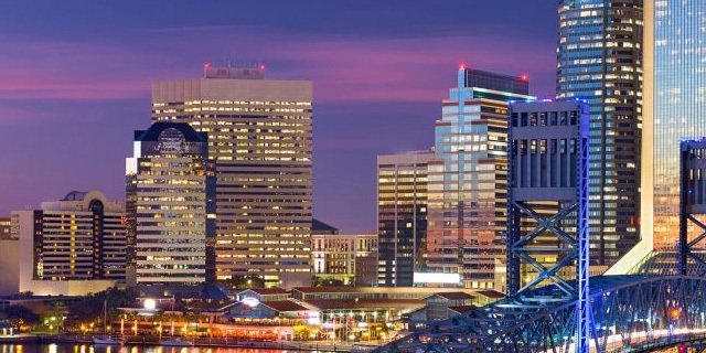 Jacksonville skyline at dusk with illuminated high-rise buildings, colorful evening sky, and bridge glowing in blue
