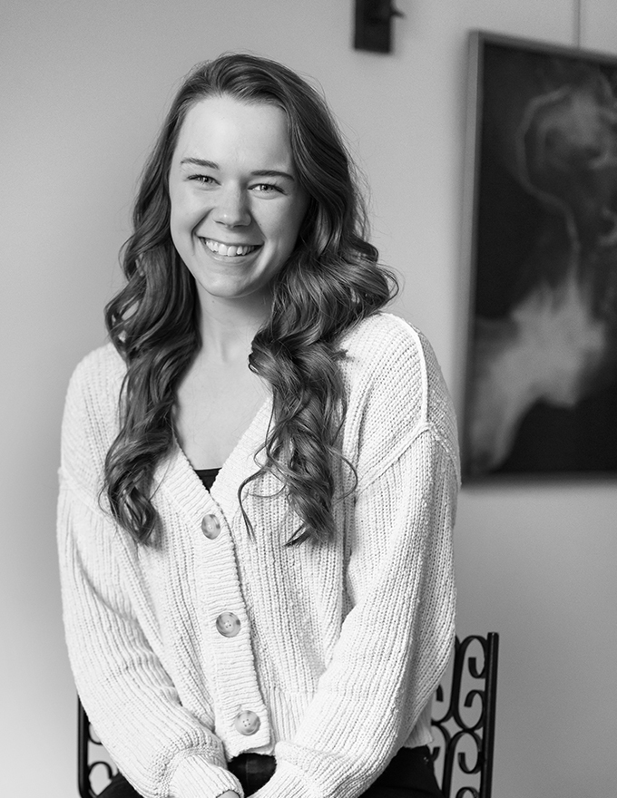 Black and white portrait of young woman smiling, wearing knit cardigan, long wavy hair, seated indoors near artwork