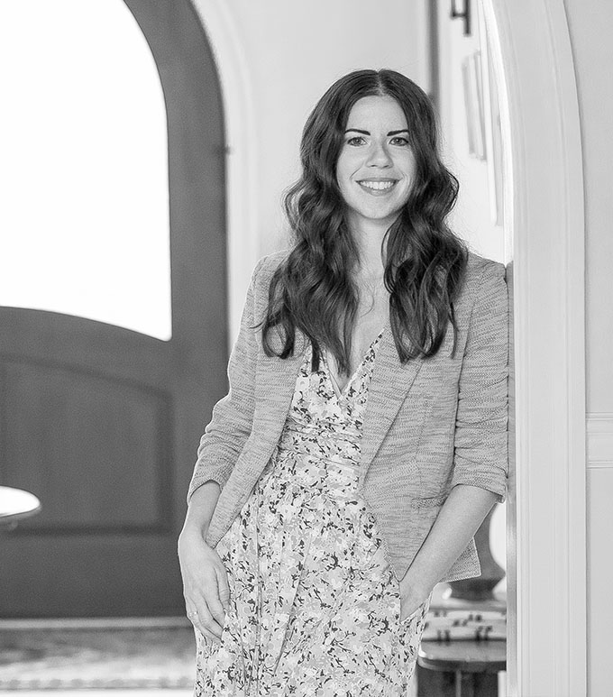Black and white portrait of woman in floral dress and blazer, smiling warmly while standing in arched doorway