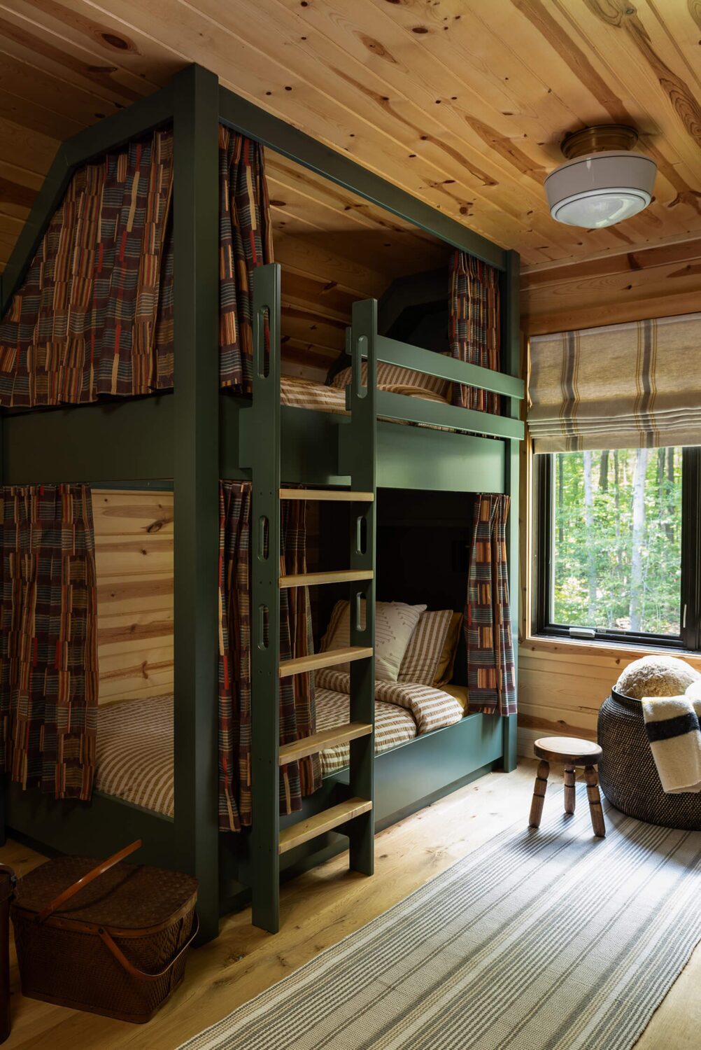 Cozy wooden cabin bedroom with built-in bunk beds, striped bedding, plaid curtains, and a window view of green forest.