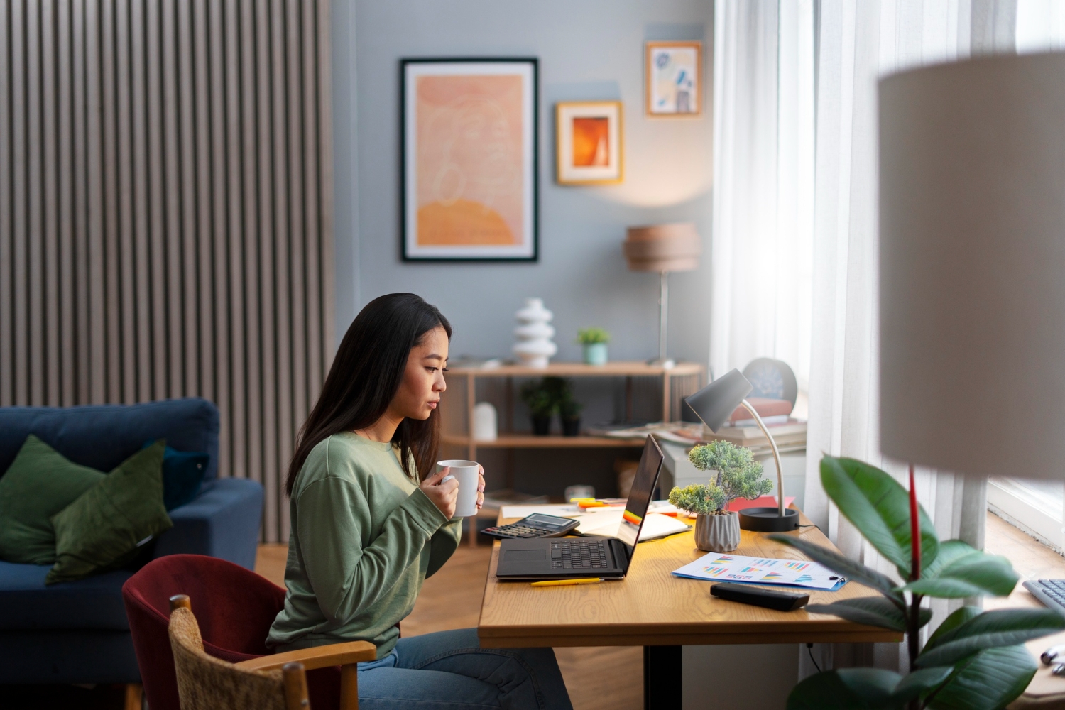 Young woman working from home at desk with laptop, coffee mug in hand, surrounded by plants, charts, and cozy decor.
