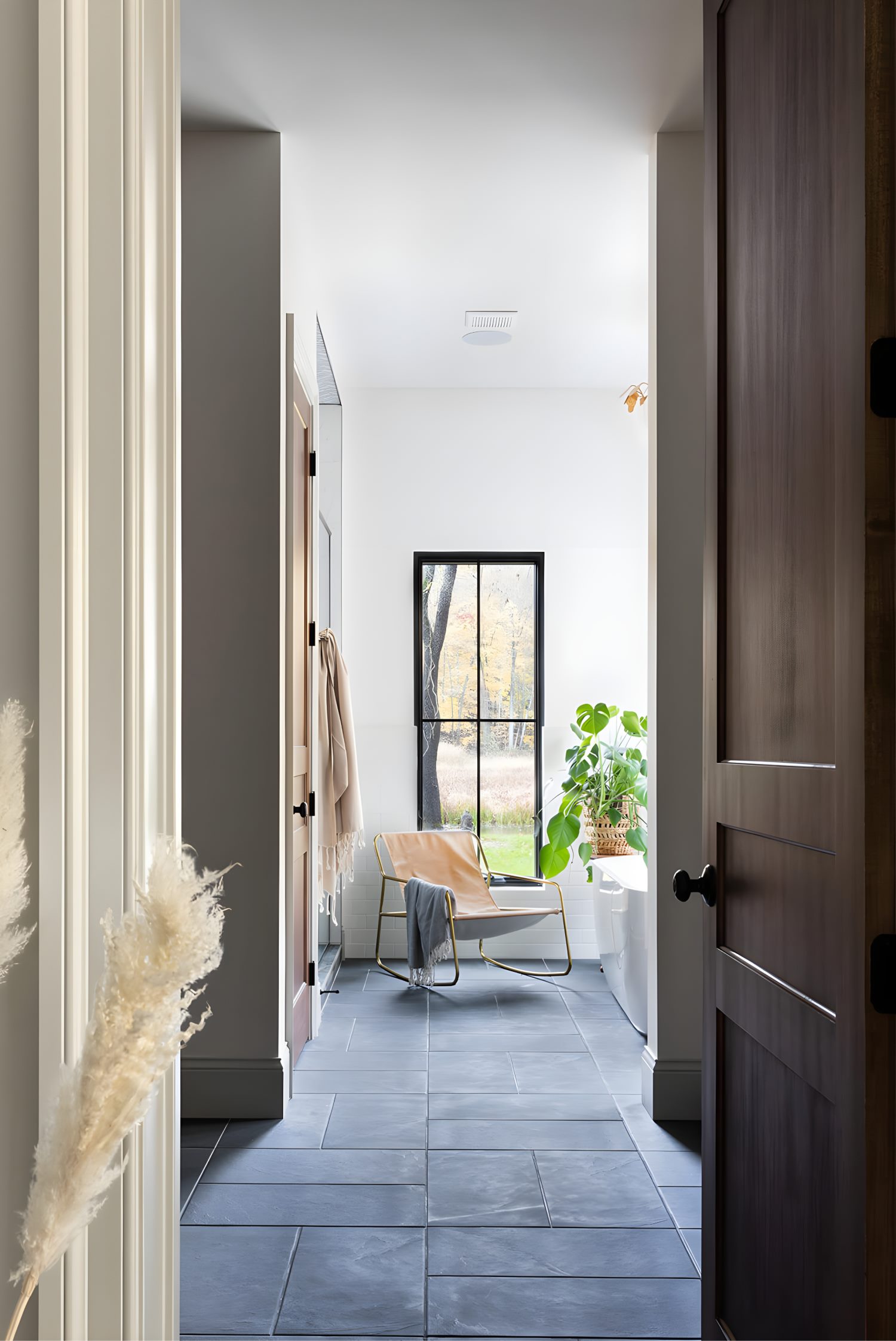 Bright bathroom with freestanding tub, large window, leafy plant, and elegant chandelier creating a relaxing spa-like retreat.