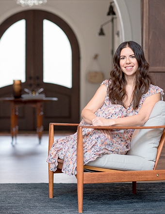Woman in floral dress sitting on a wooden chair indoors, smiling with a bright background and large wooden doors behind.