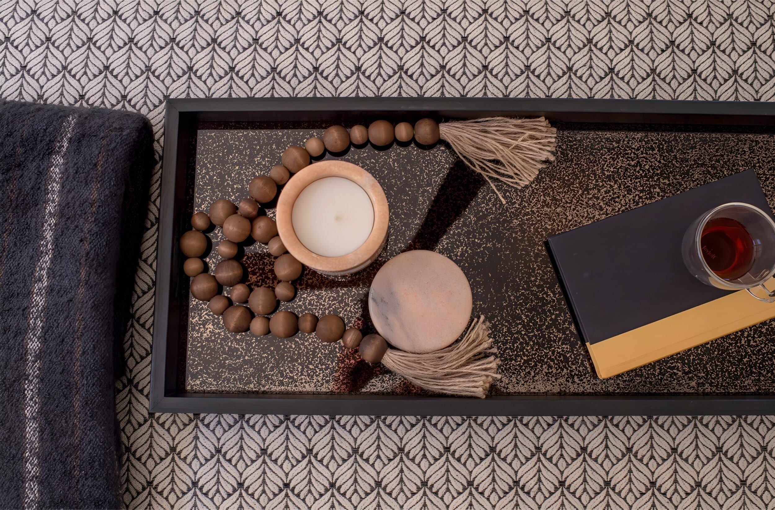 Decorative tray on patterned bench with candle, wooden beads, book, and glass of tea arranged neatly in a cozy setting