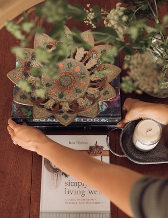 Overhead view of hands arranging a decorative flower-shaped piece on books with a candle, plants, and a lifestyle guide.