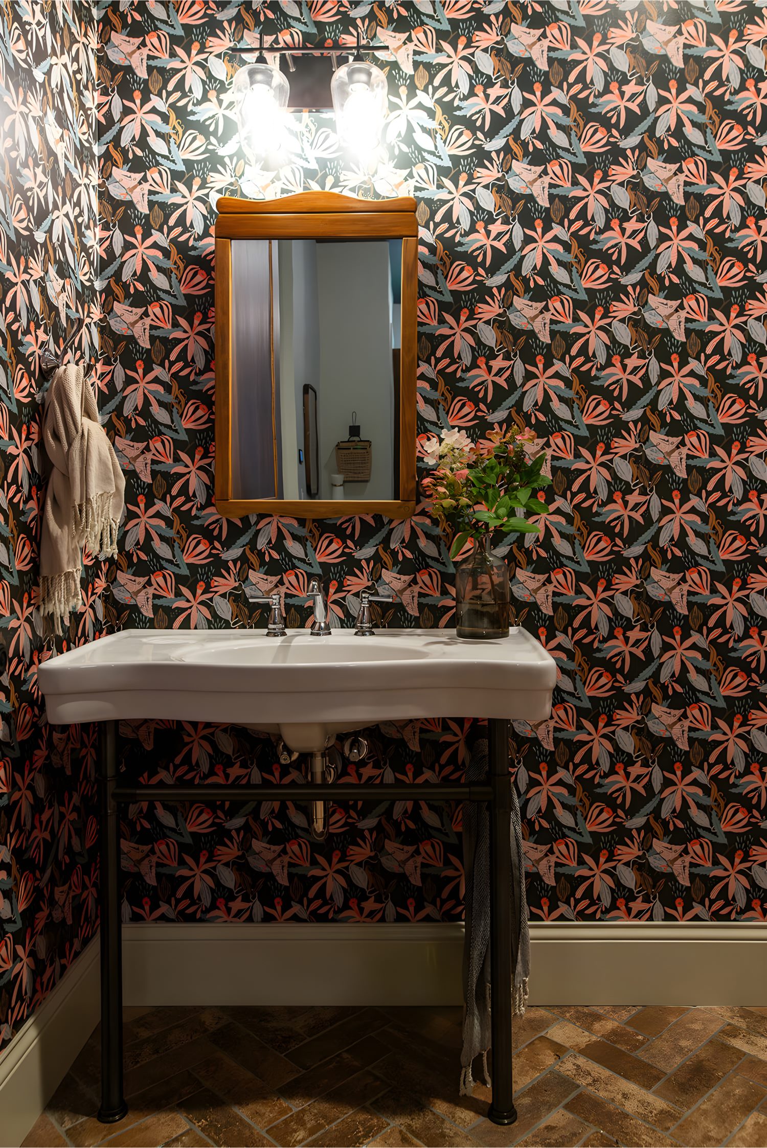 Powder room with bold floral wallpaper, wall mirror, white sink, and vase of fresh flowers as accent.