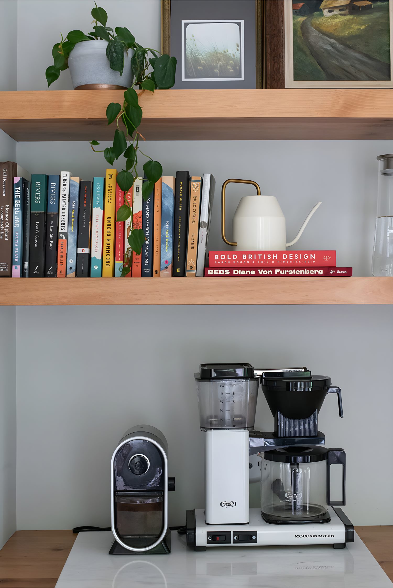 Bookshelf with stacked books, framed art, trailing plant, and coffee machines below on countertop.