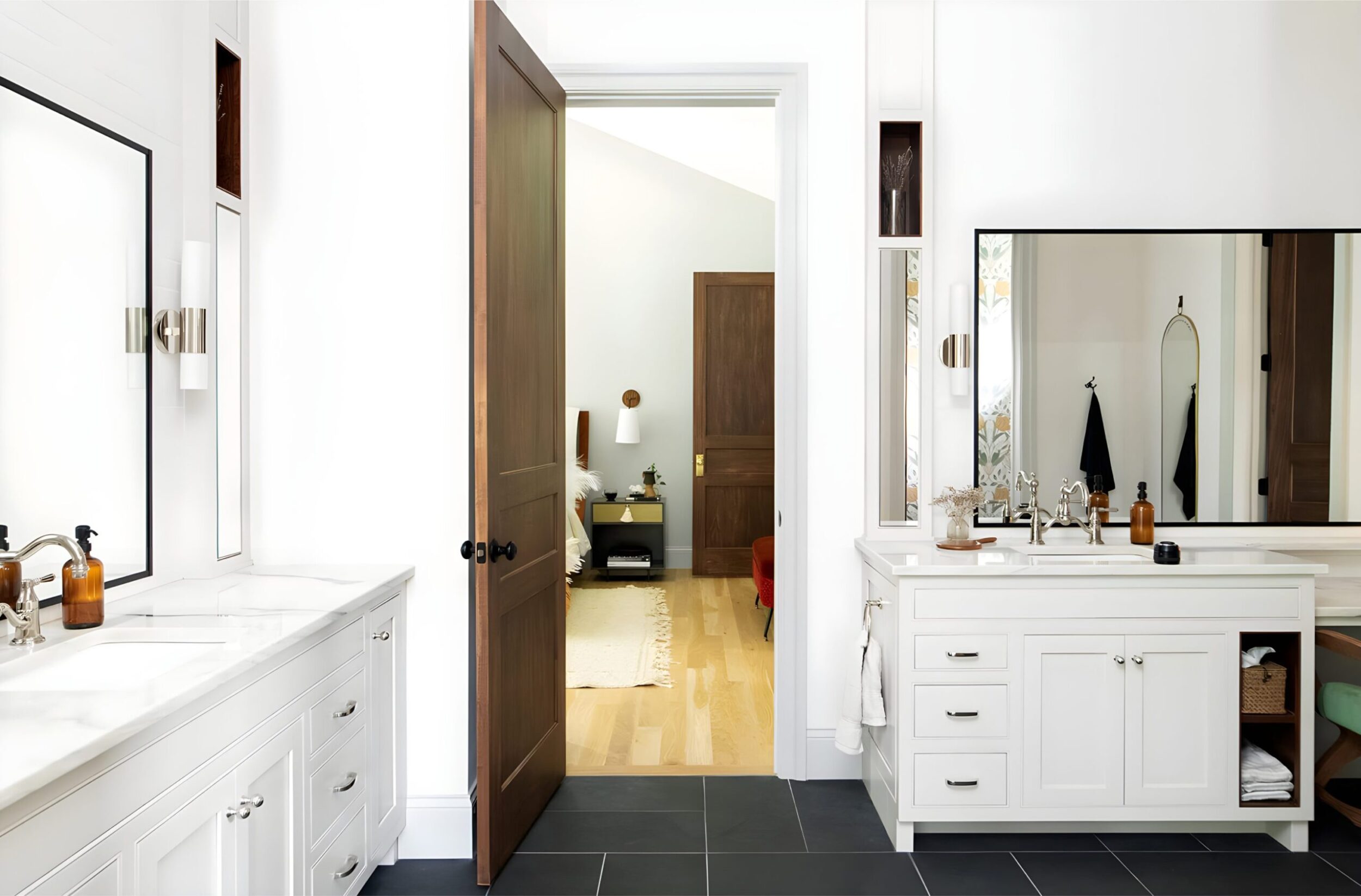 White bathroom with dual vanities, wood doors, large mirrors, and black tile floor leading to bedroom.