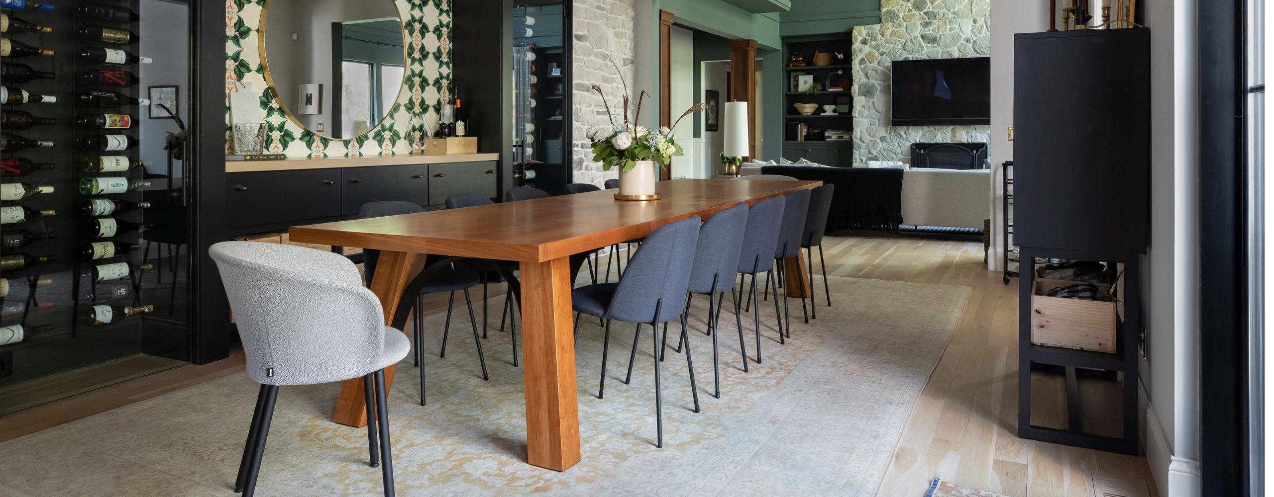 Dining room with wood table, upholstered chairs, stone wall, wine display, round mirror, and view into cozy living area