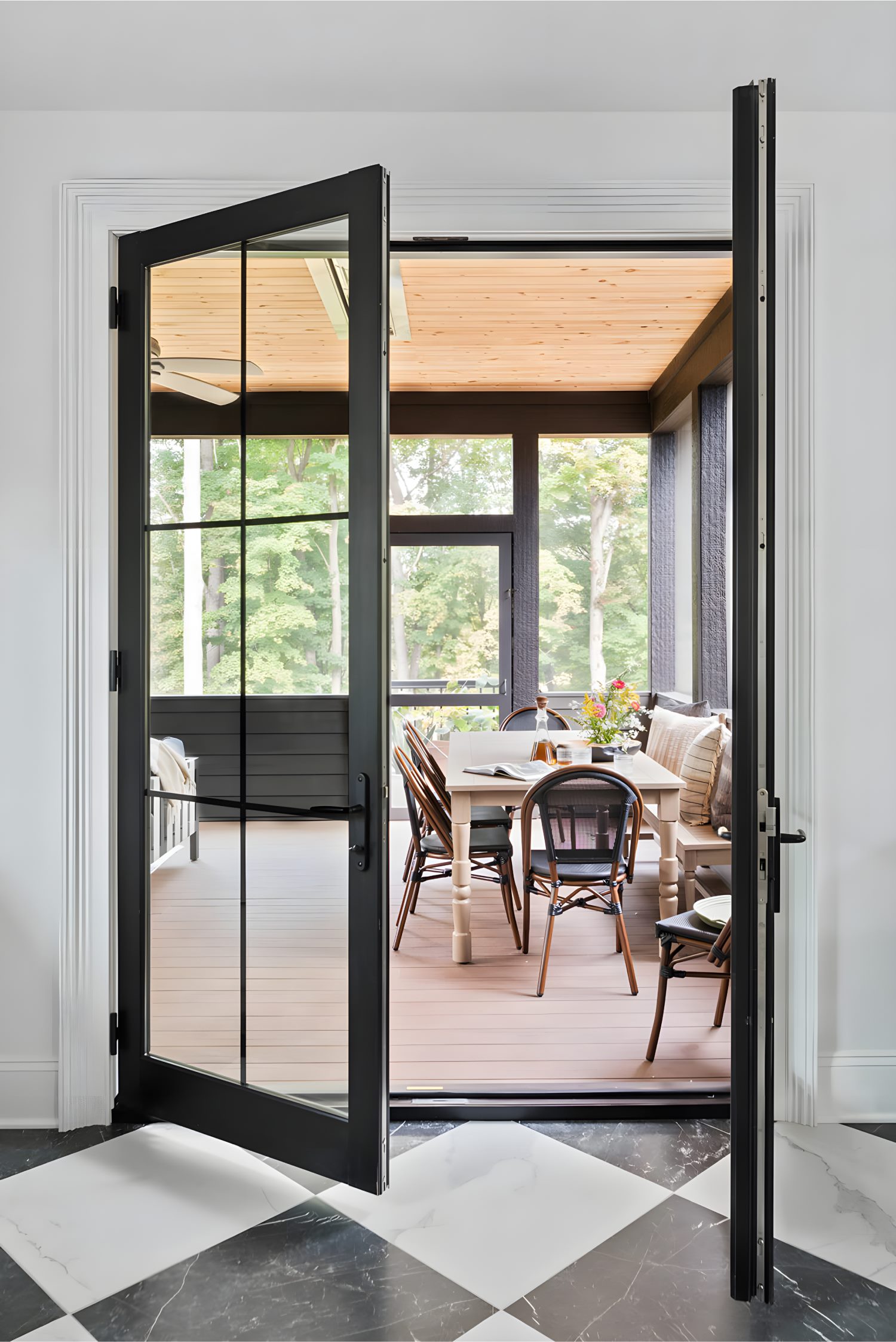 Black-framed glass door opening to porch dining area with wood table, wicker chairs, and soft light through large windows