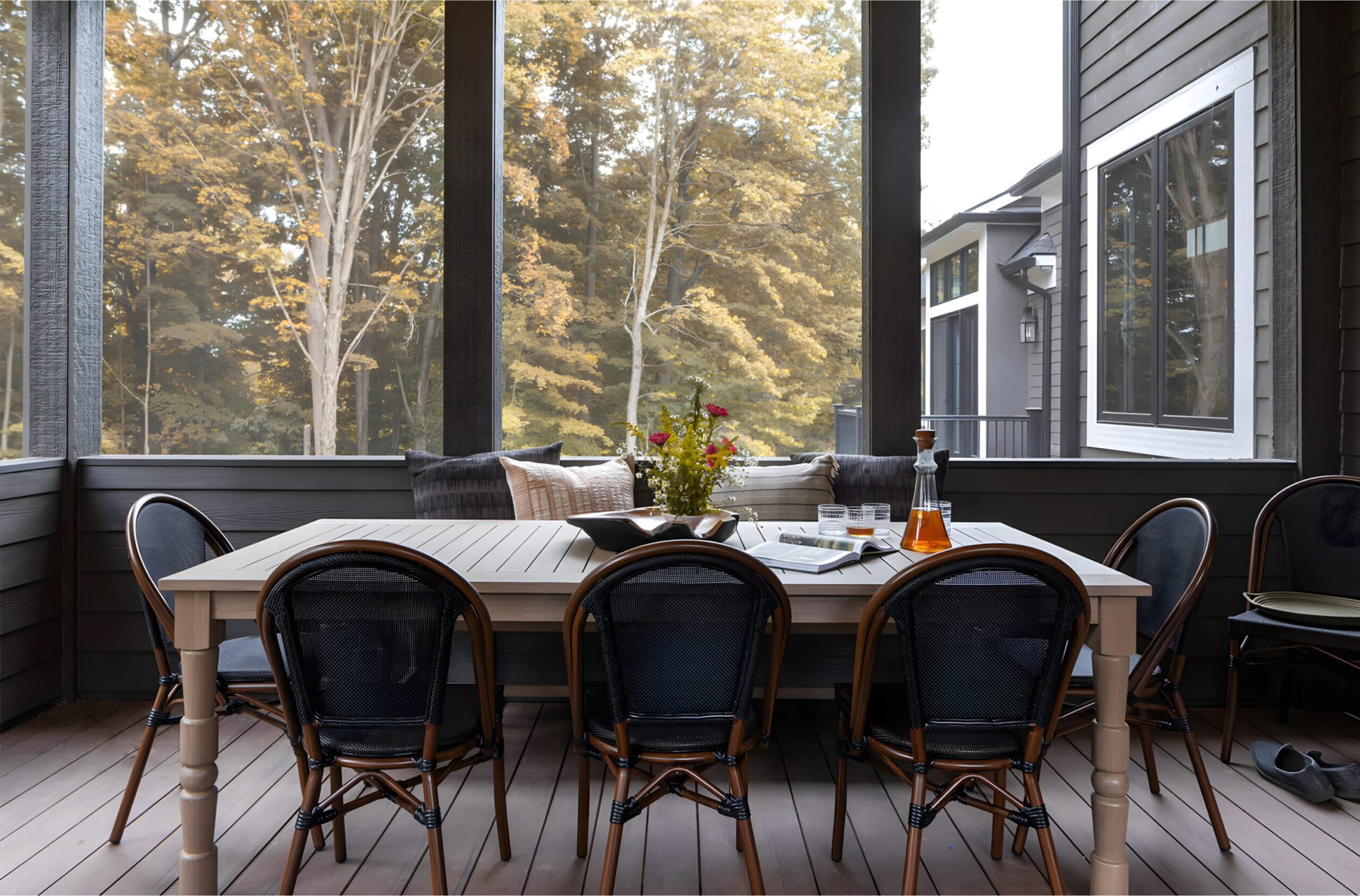 Screened-in porch with wooden dining table, black chairs, throw pillows, and glassware against autumn tree backdrop