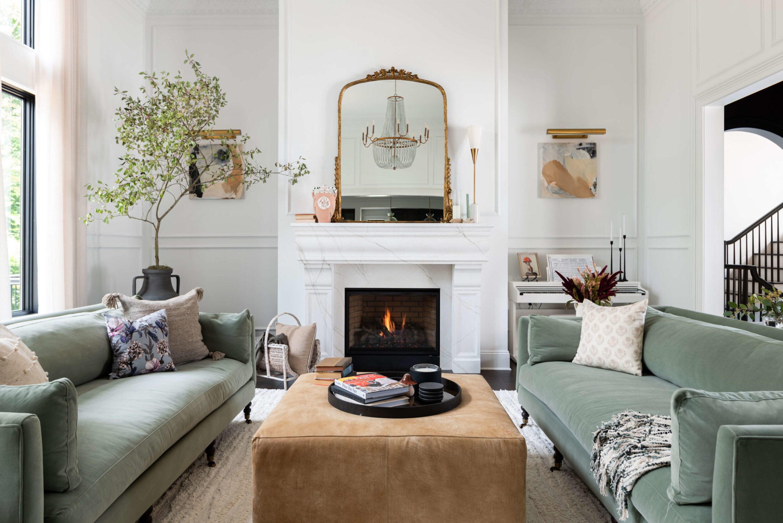 Sophisticated living room with twin sage green sofas, tan ottoman, white fireplace and large gold framed mirror
