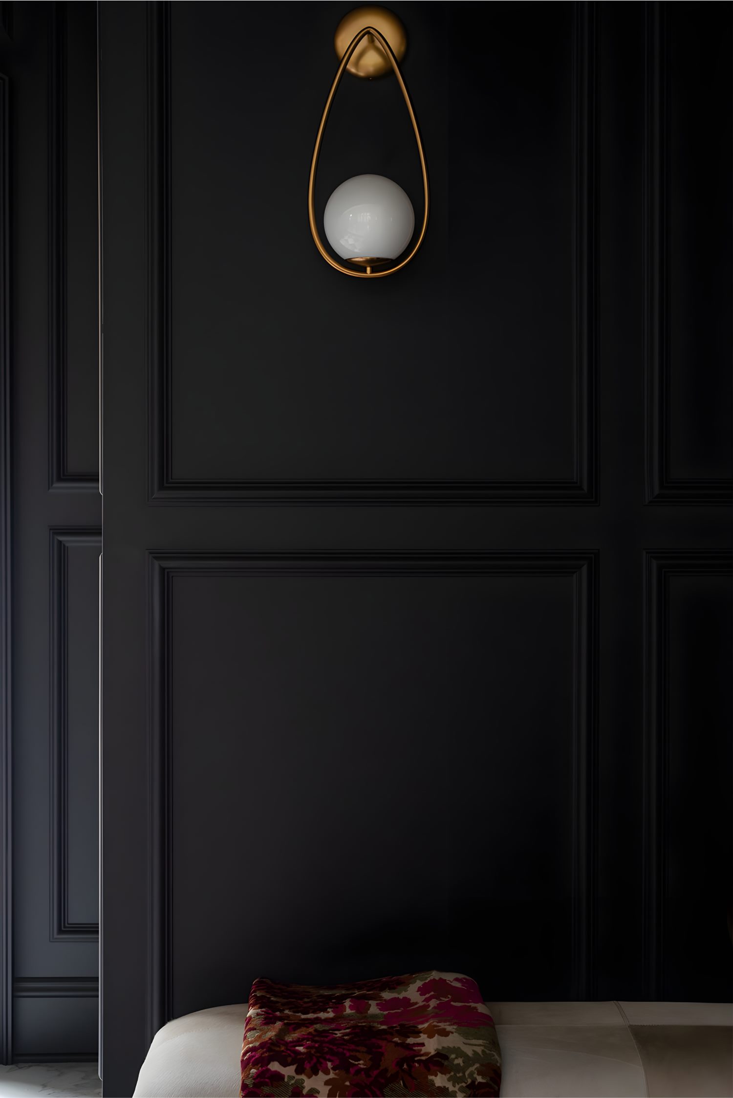 Black paneled wall with modern gold sconce holding a white globe light, above a bench draped with patterned throw