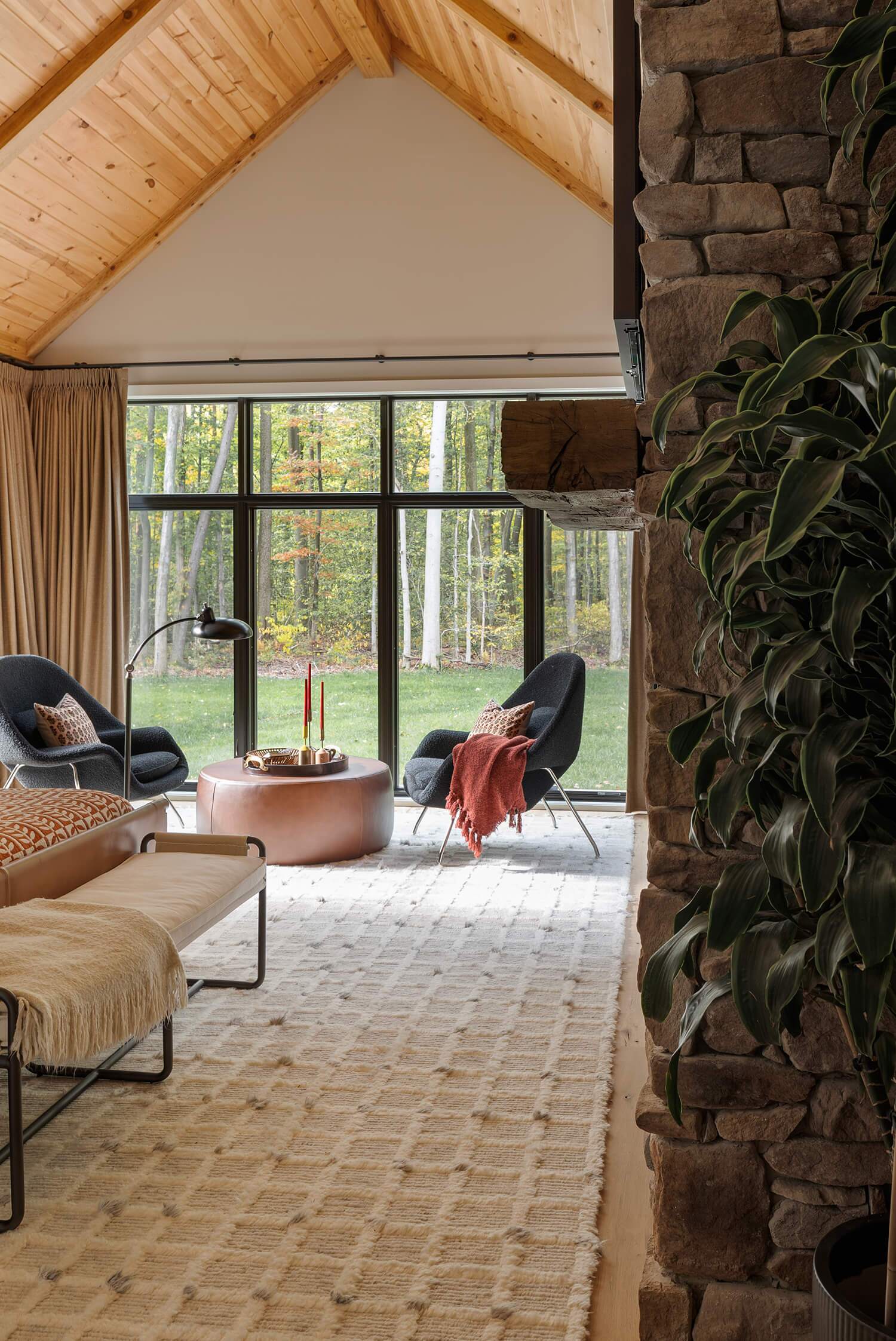 Living area with vaulted wood ceiling, stone fireplace, modern chairs, and large windows overlooking autumn landscape