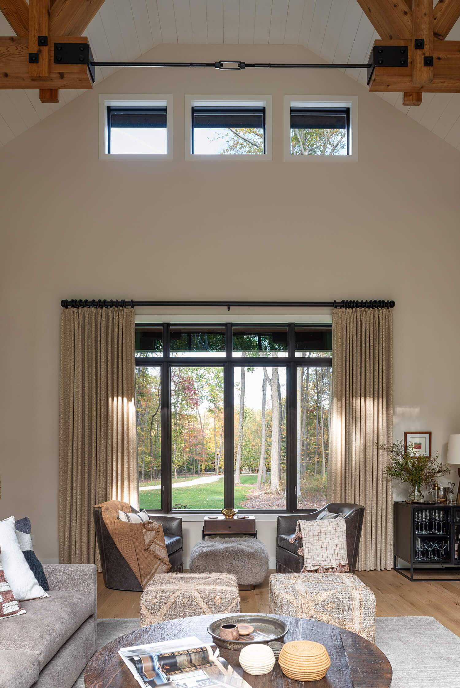 Bright living room with vaulted ceiling, tall windows, neutral sofas, textured ottomans, and autumn forest views