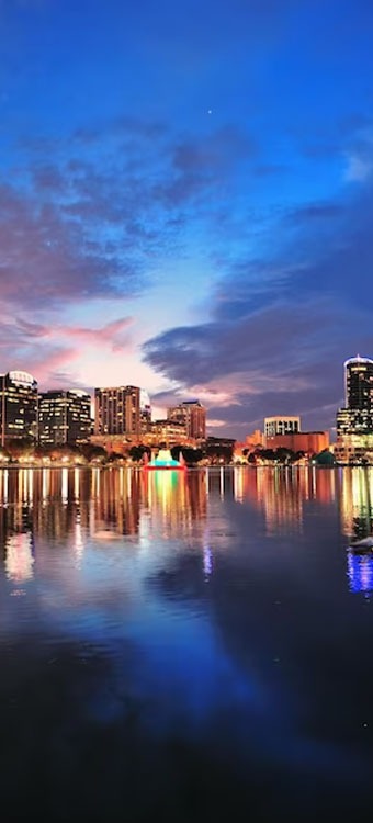City skyline at dusk reflecting on lake with fountain lit in colors, tall buildings glowing under vibrant evening sky