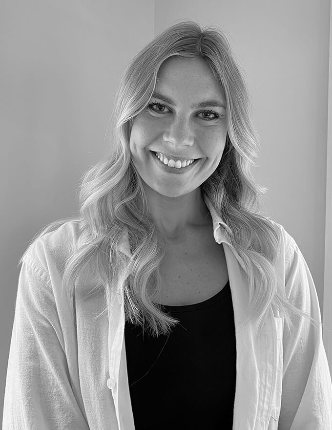 Emilie Designer, Black and white portrait of a smiling woman with wavy blonde hair, wearing a light shirt over a dark top, against a plain wall.