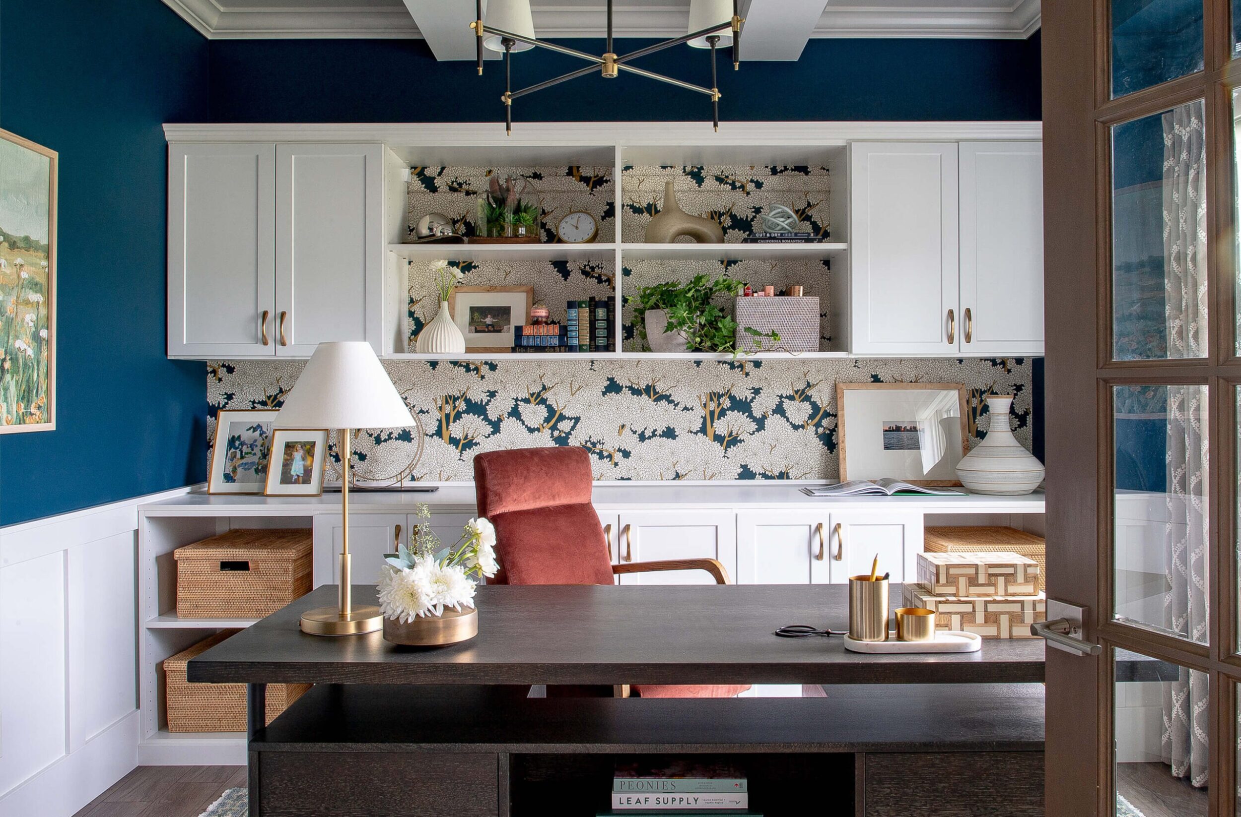 Modern office with bold blue walls, white cabinetry, floral wallpaper, dark desk, and coral chair styled with gold accents.