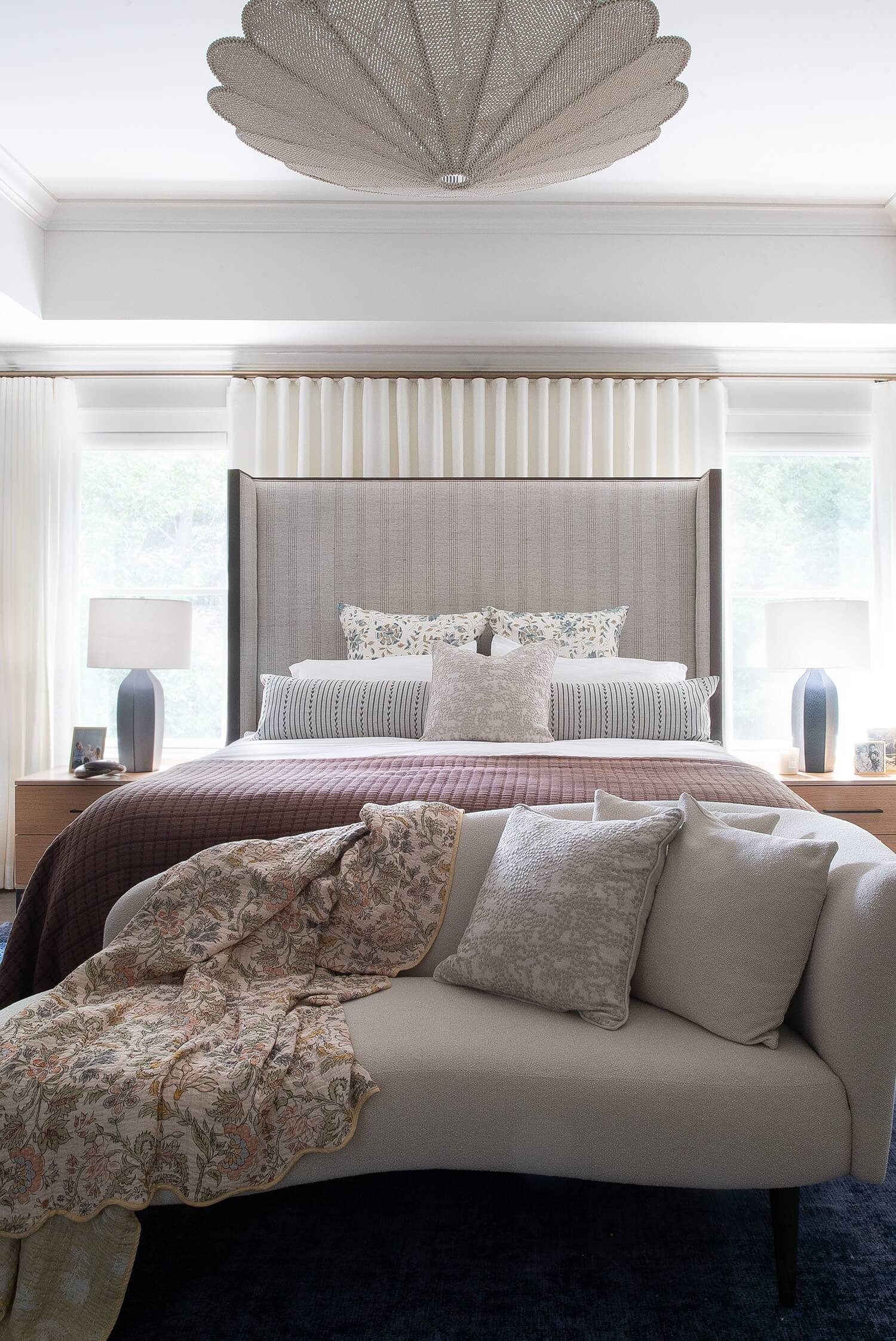 Bedroom featuring tall upholstered bed, patterned pillows, plum quilt, and accent loveseat, styled with modern lighting.