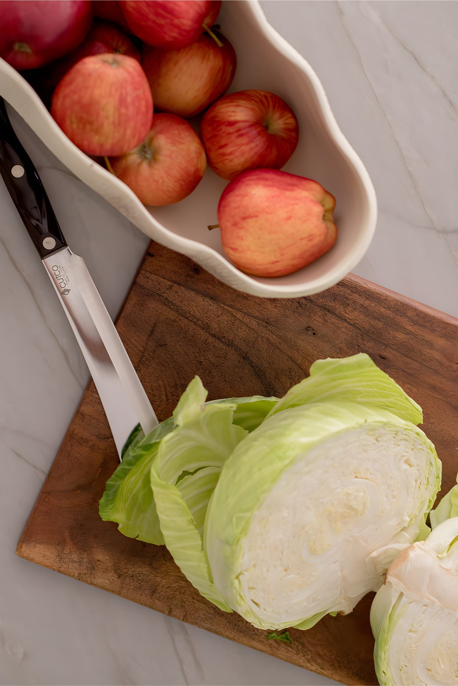 Close-up of cutting board with halved cabbage, apples in ceramic bowl, and knife