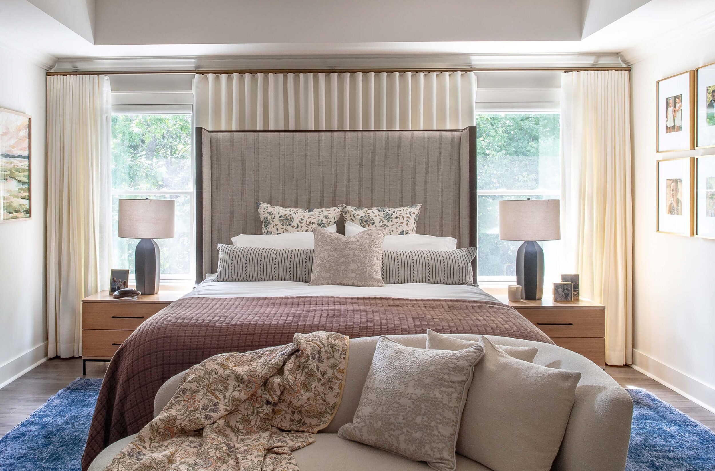 Bright bedroom with upholstered headboard, bedside lamps, floral artwork, and neutral bedding accented by plum quilt.