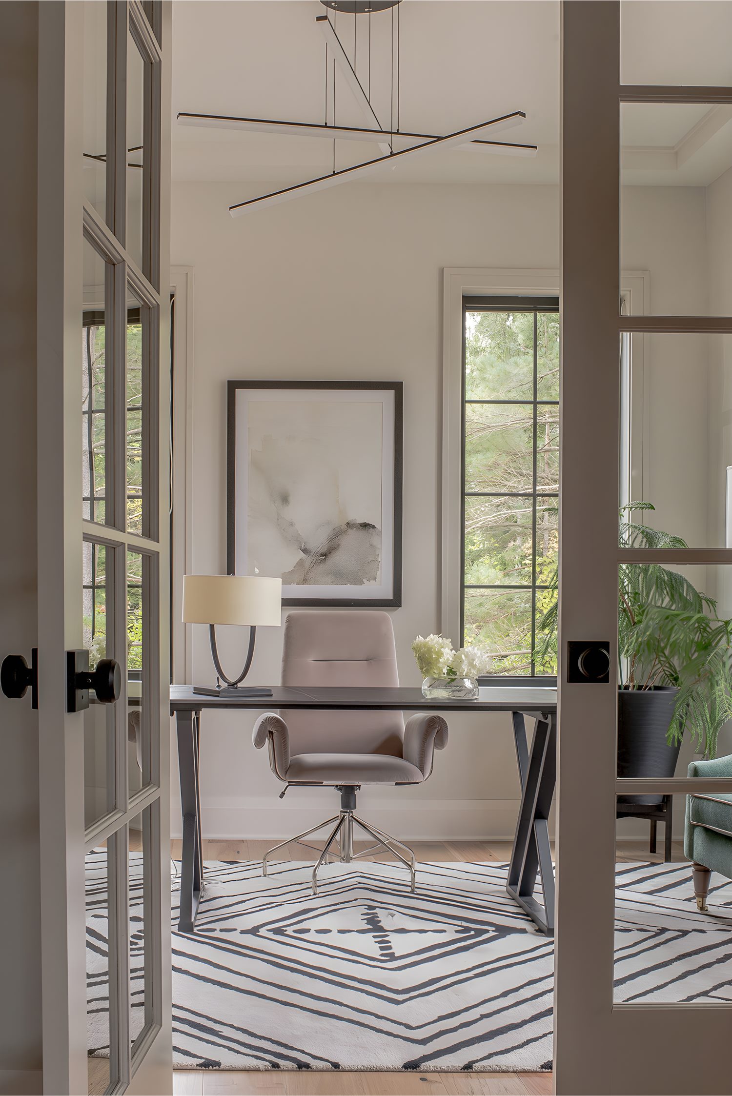 Home office with modern desk, blush swivel chair, large windows, abstract wall art, and geometric rug