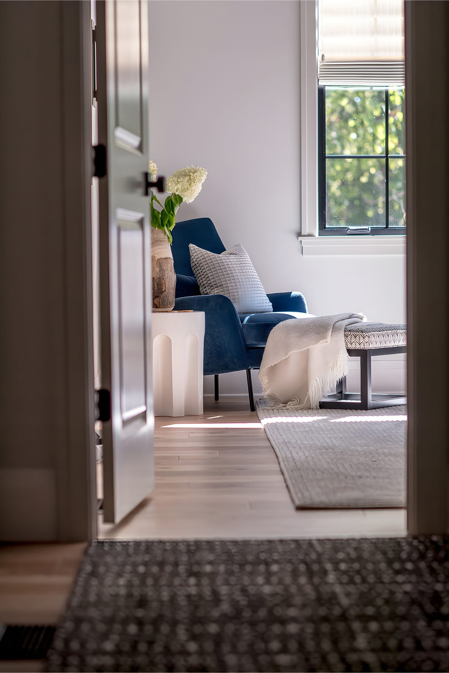 Cozy bedroom corner with blue armchair, white pillow, side table, and natural light from window with greenery view