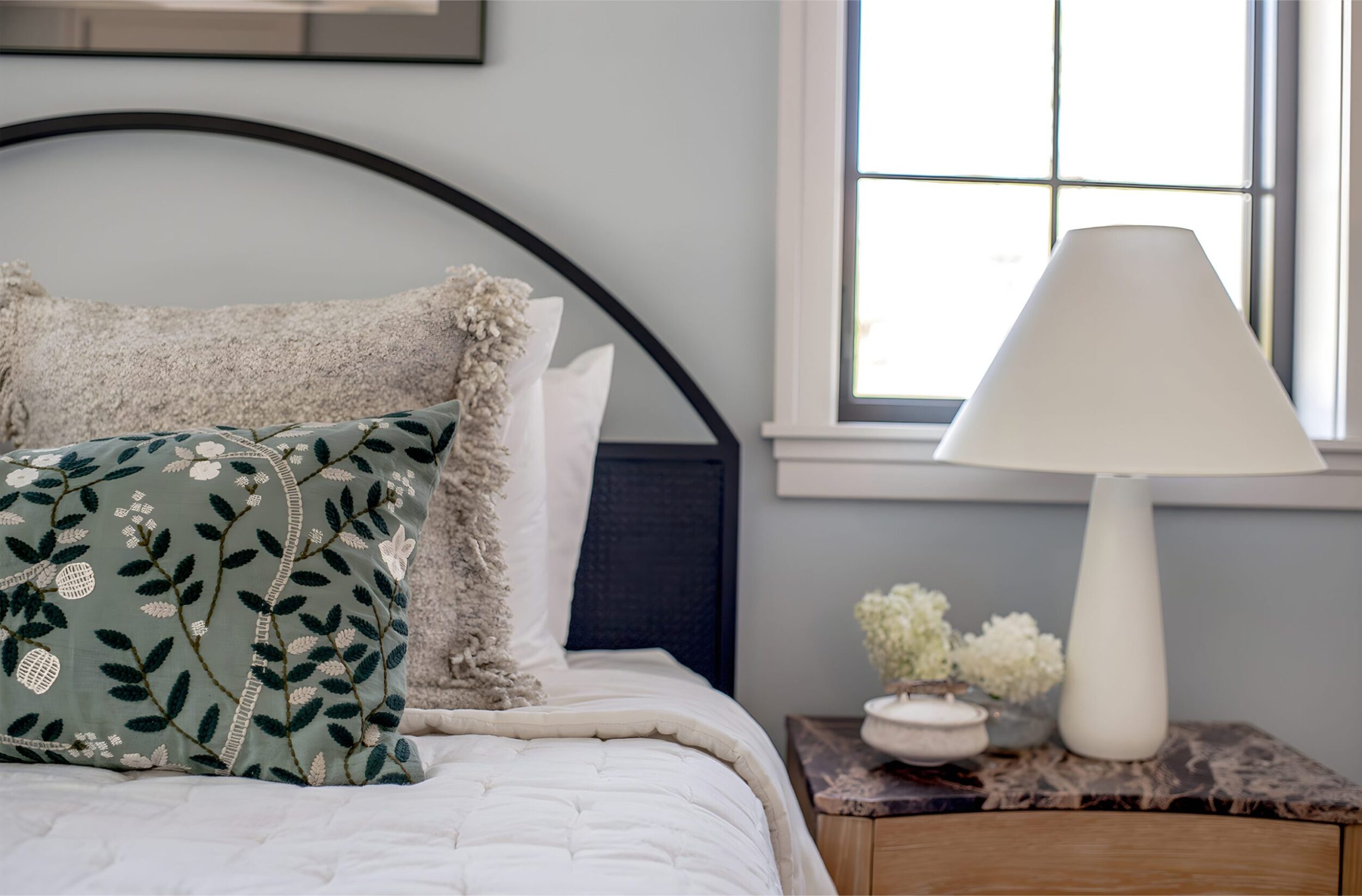 Cozy bedroom corner with textured pillows, floral accent cushion, and white lamp on nightstand beside window.