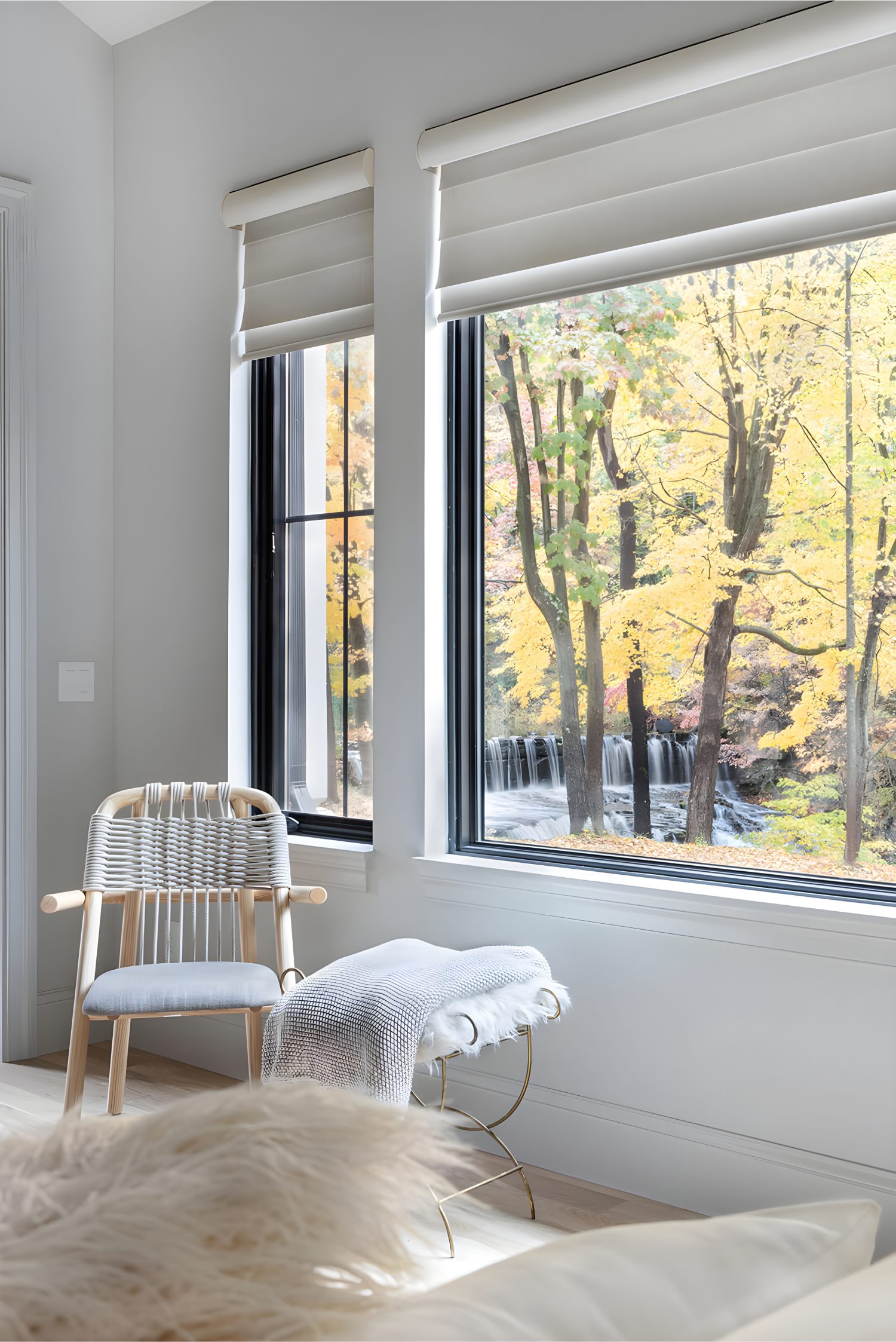 Bright bedroom corner with large black-framed windows, white blinds, and cozy chair overlooking a scenic waterfall and autumn trees