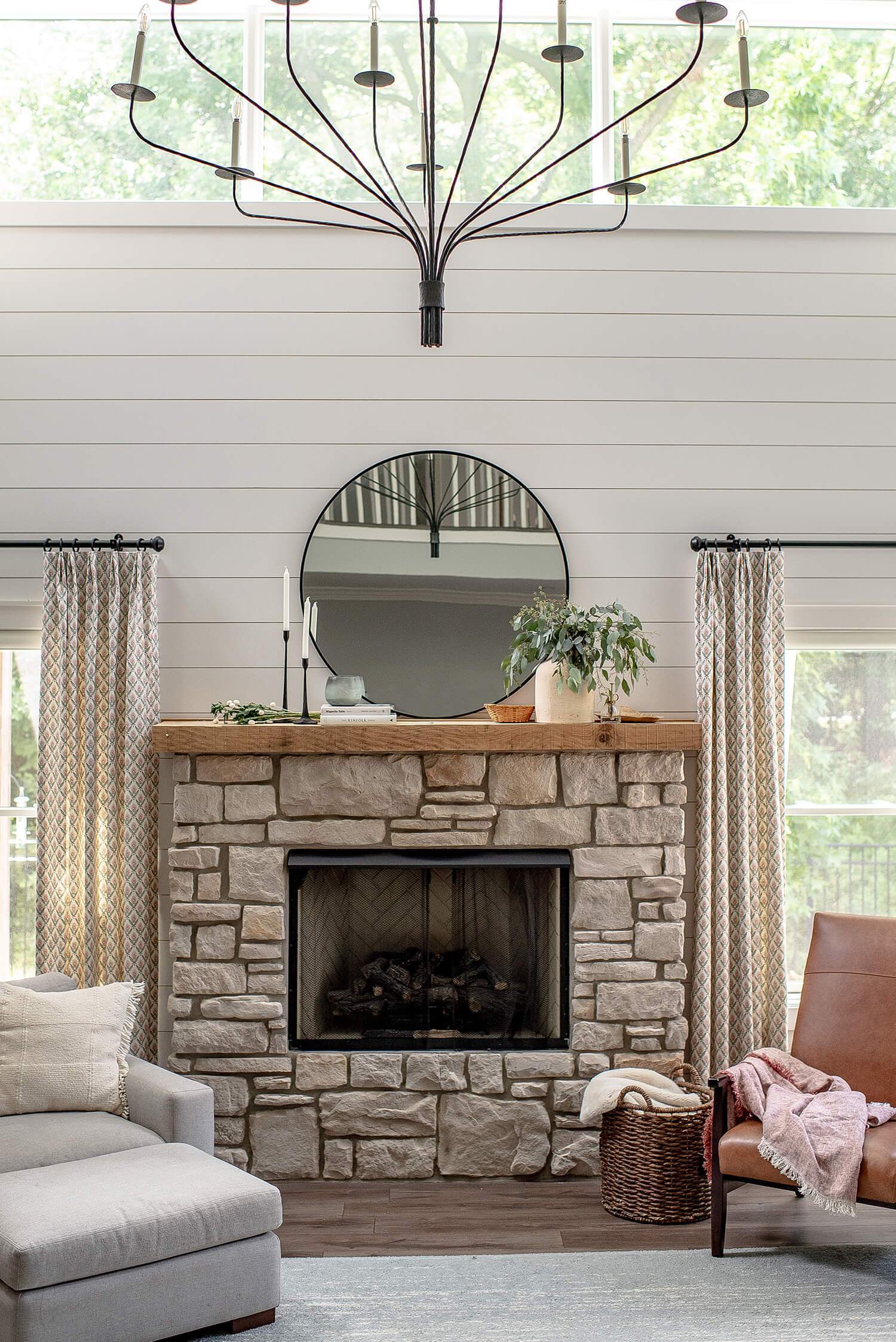 Cozy stone fireplace with wood mantel, round mirror, plants, and seating styled with soft textiles and neutral decor.