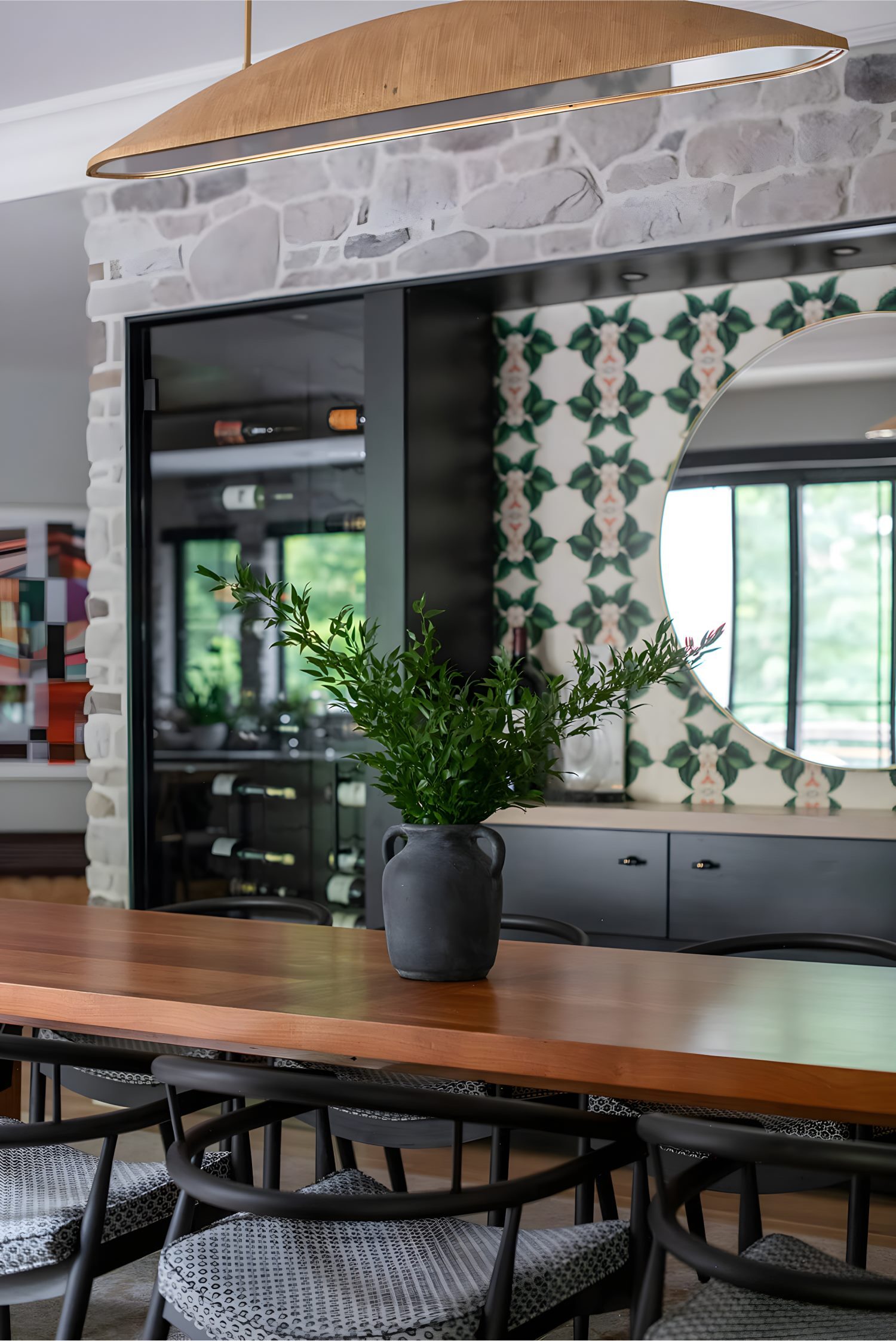 Modern dining area with wood table, black chairs, large mirror, and vase centerpiece against patterned wall.