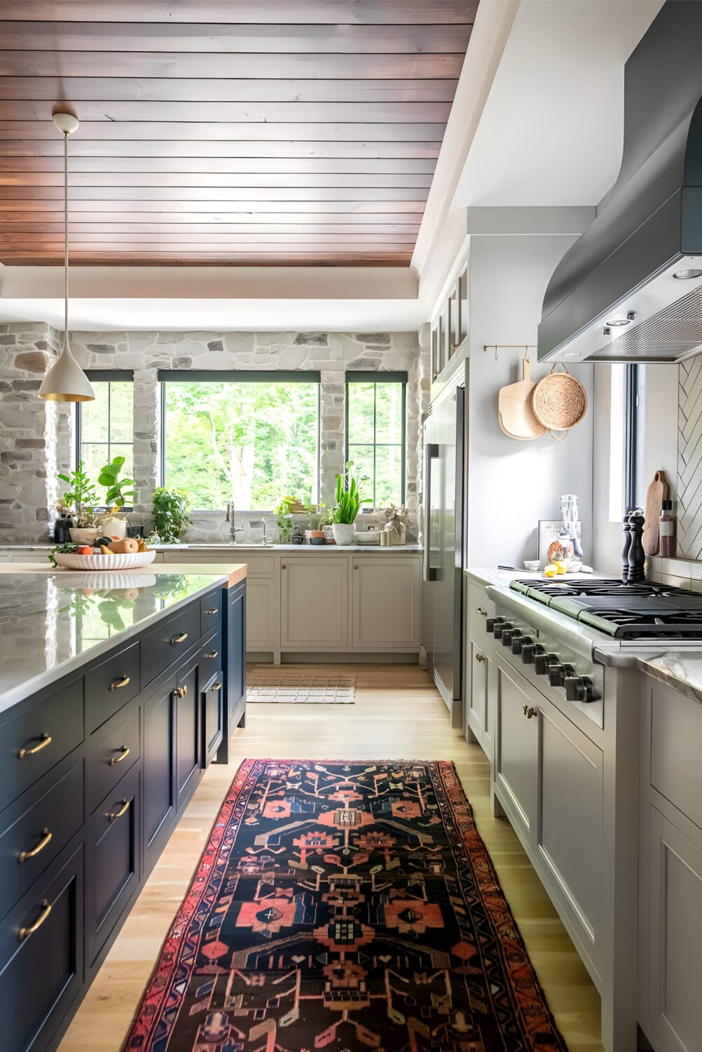 Bright kitchen with stone walls, wood ceiling, dark island, patterned rug, and large windows with greenery view.