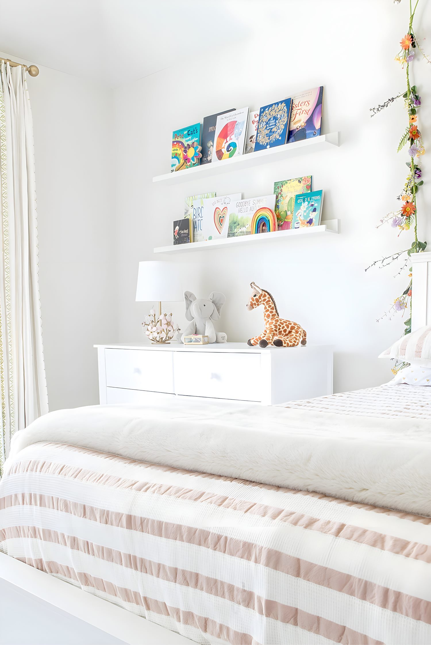 White kids’ room with book display shelves above dresser, stuffed giraffe toy, and floral garland accent on wall