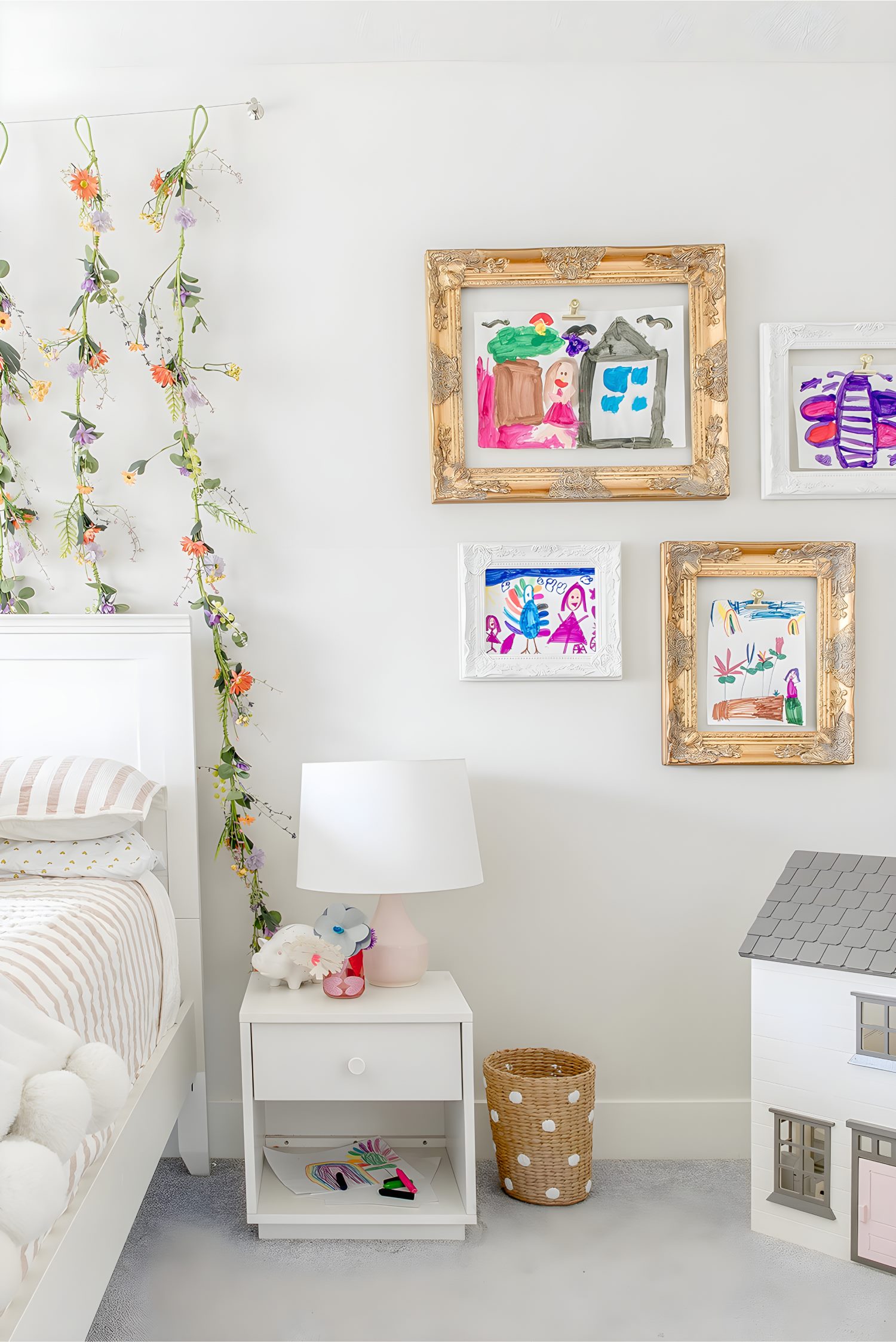 Gallery wall of framed children’s artwork in gold and white frames beside bed with floral garland accent
