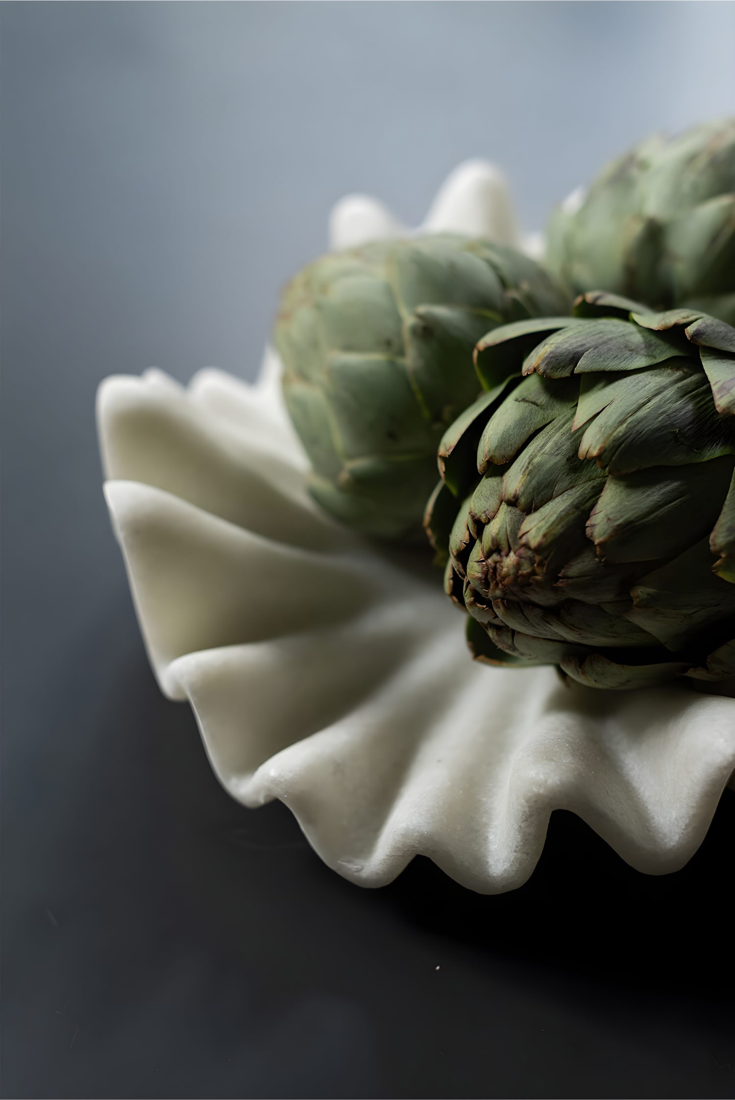 Close-up of sculpted white ceramic bowl with green artichokes, placed on dark countertop in modern kitchen setting