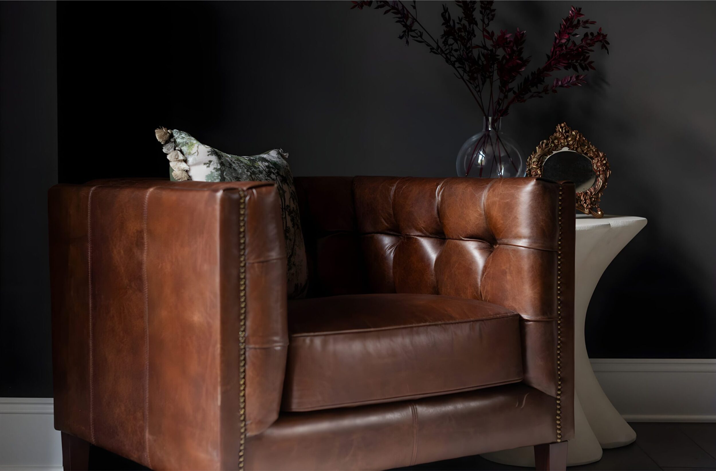 Brown tufted leather armchair with patterned pillow beside a vase with branches and ornate gold decorative mirror