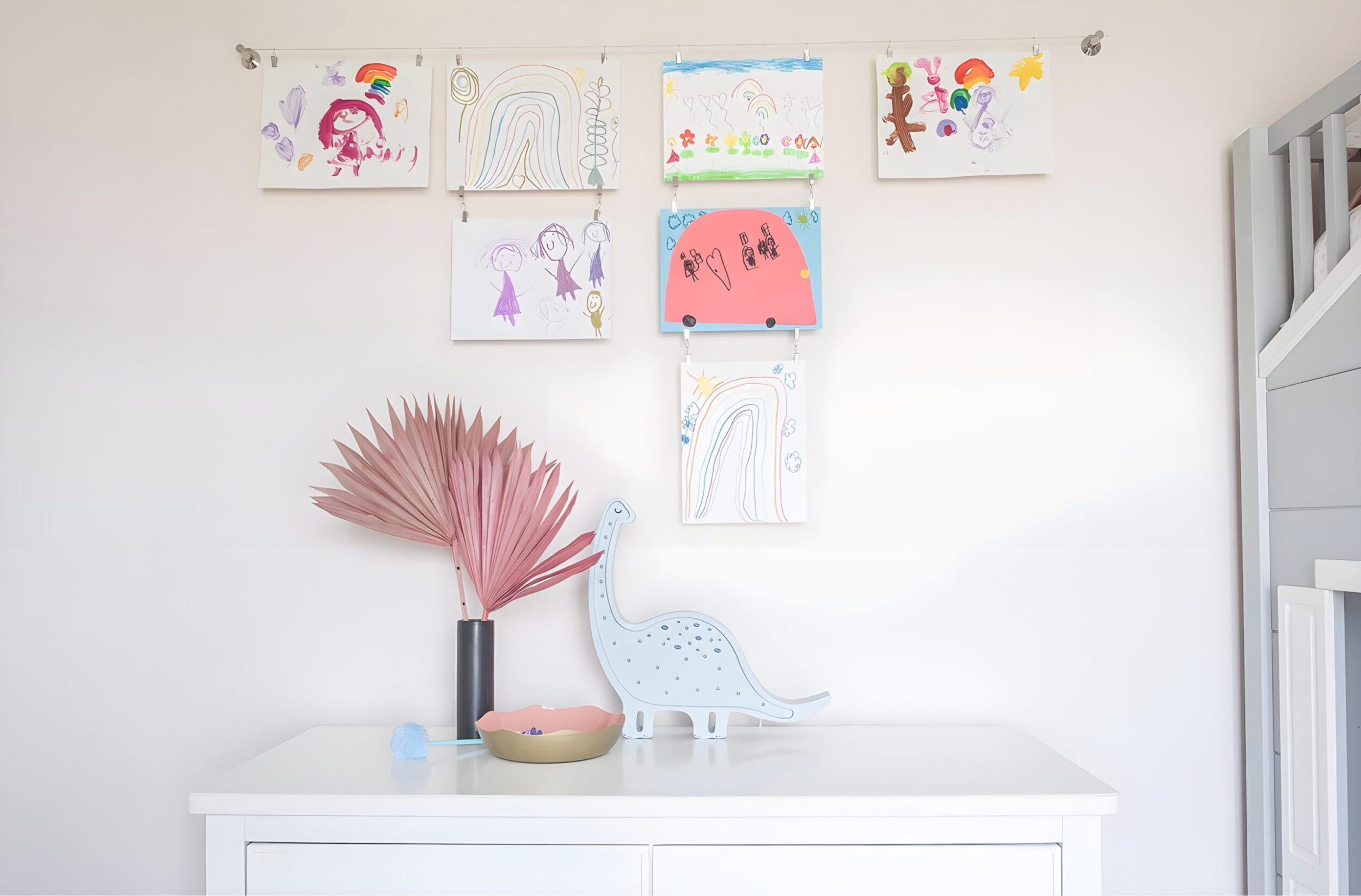 Children’s colorful artwork hung on wall with clips above dresser, styled with pink palm leaf decor and blue dinosaur