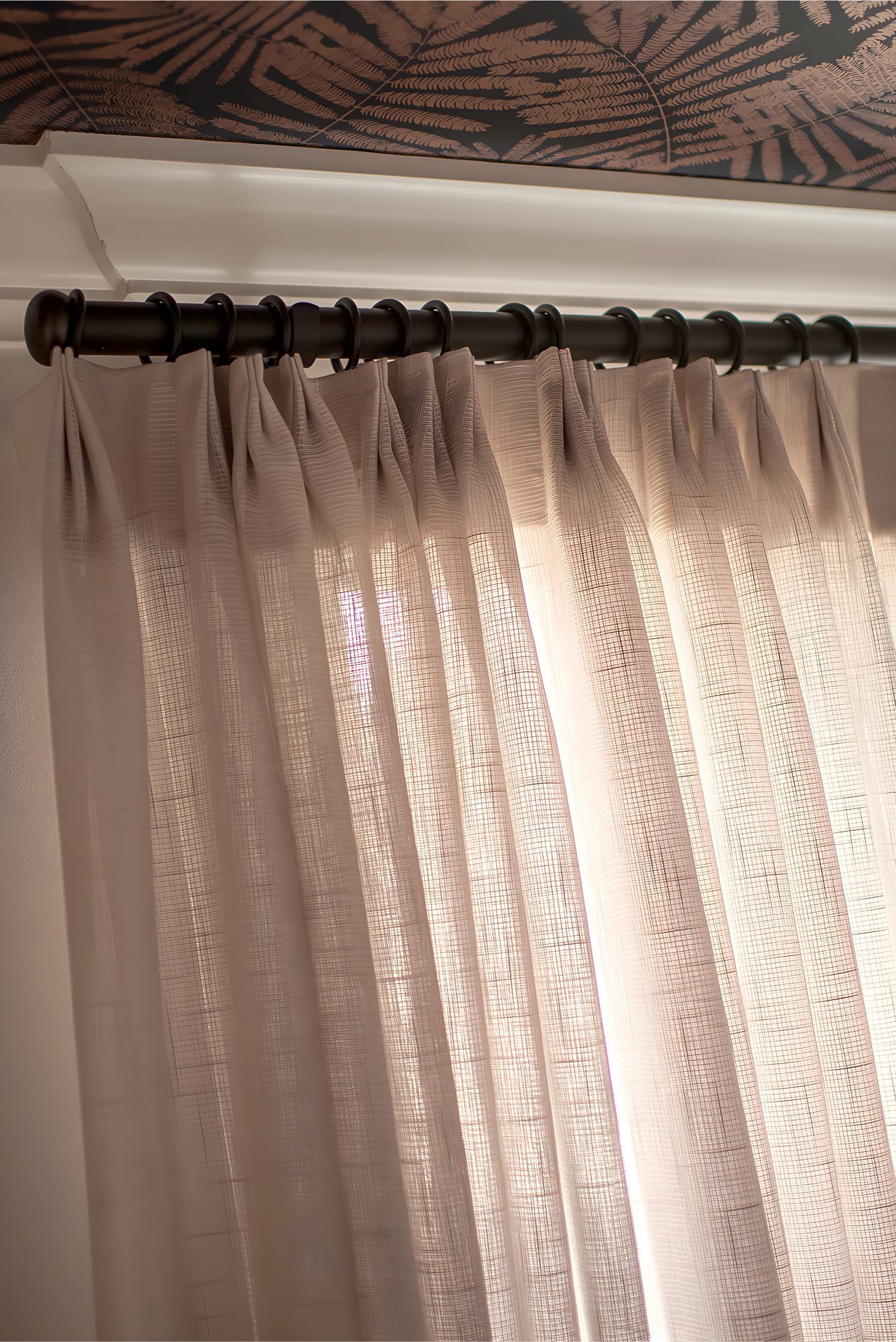 Beige sheer curtains on black curtain rod with crown molding and fern-patterned ceiling wallpaper above.