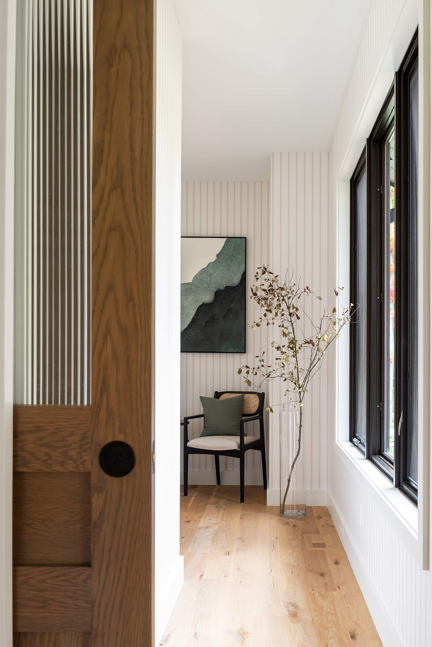 Bright hallway with wood floors, striped white wall panels, framed art, chair, and tall plant in glass vase by window