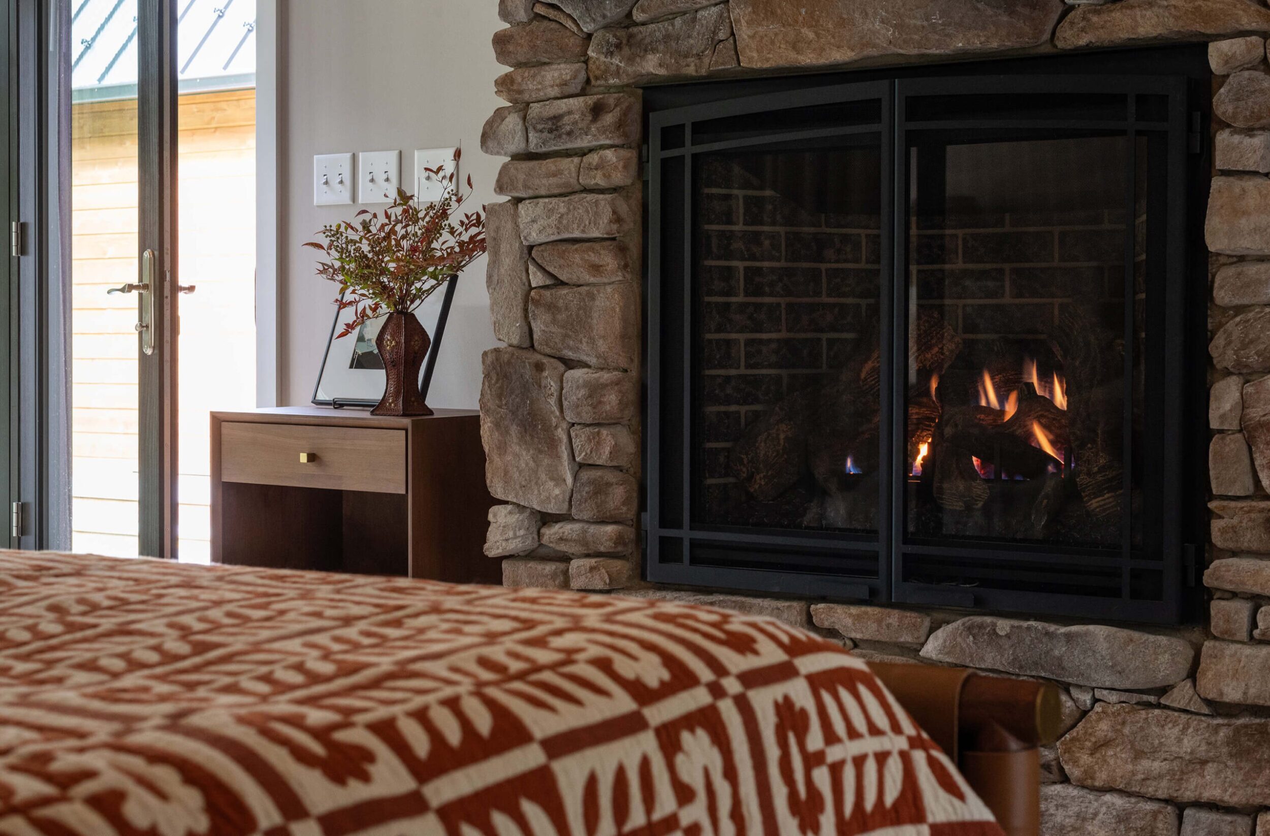 Bedroom corner with stone fireplace, wood side table, vase, and cozy bed featuring patterned red quilt and warm tones