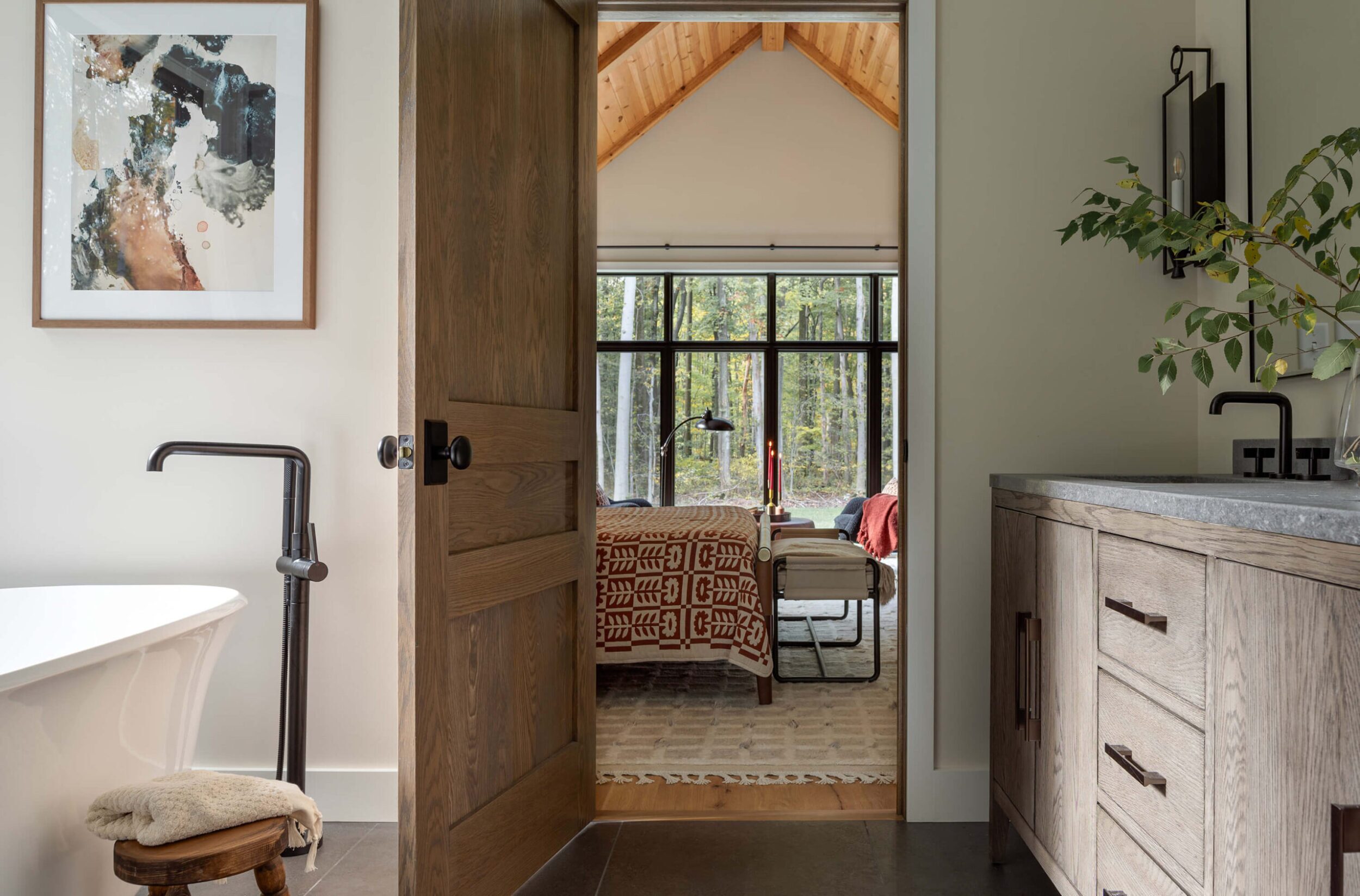 Bathroom view with freestanding tub, wooden door opening to bedroom with forest view