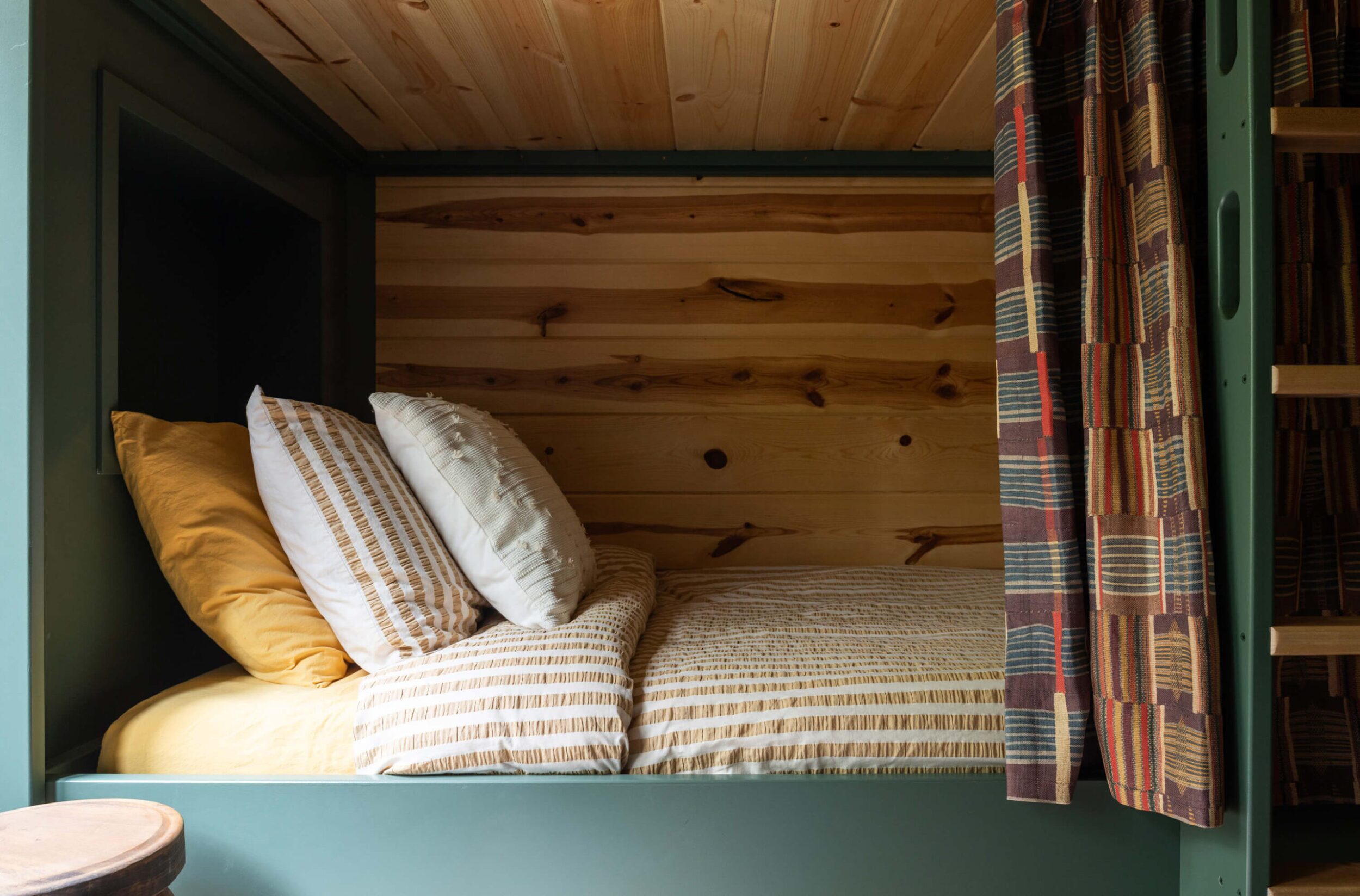 Wood-clad bunk with striped bedding, layered neutrals and mustard pillows, moss-green frame, patterned curtain, cozy ambient glow.