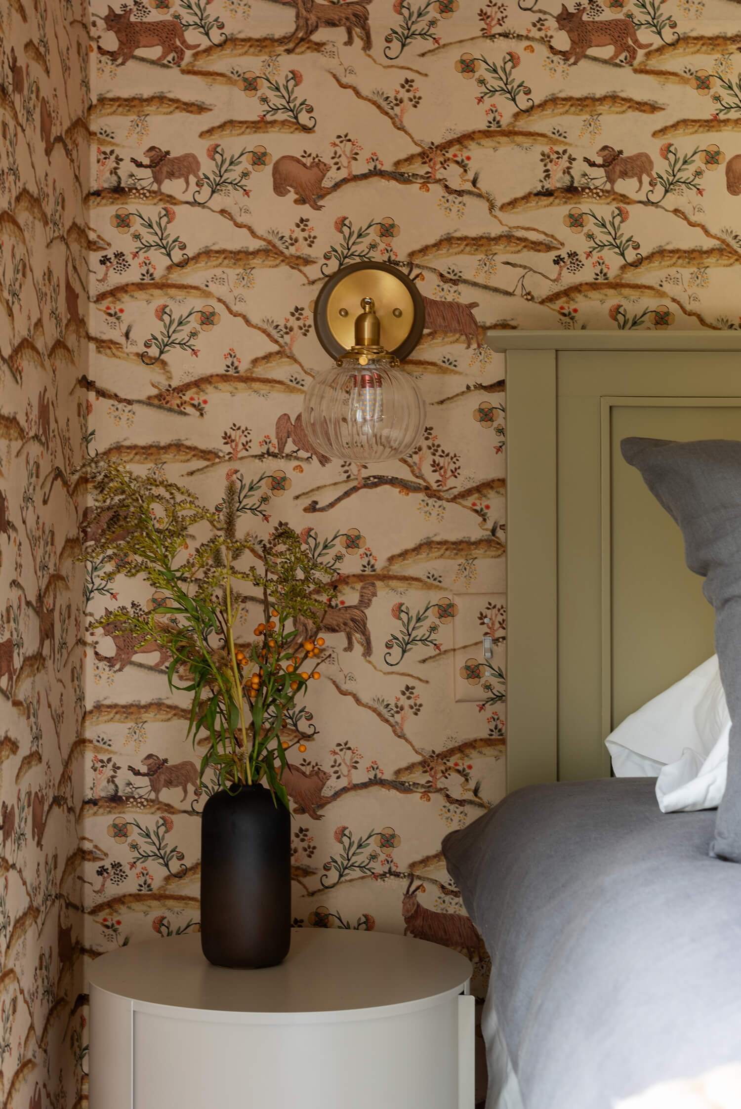 Green headboard bedroom with woodland wallpaper, black vase of flowers, and glass sconce