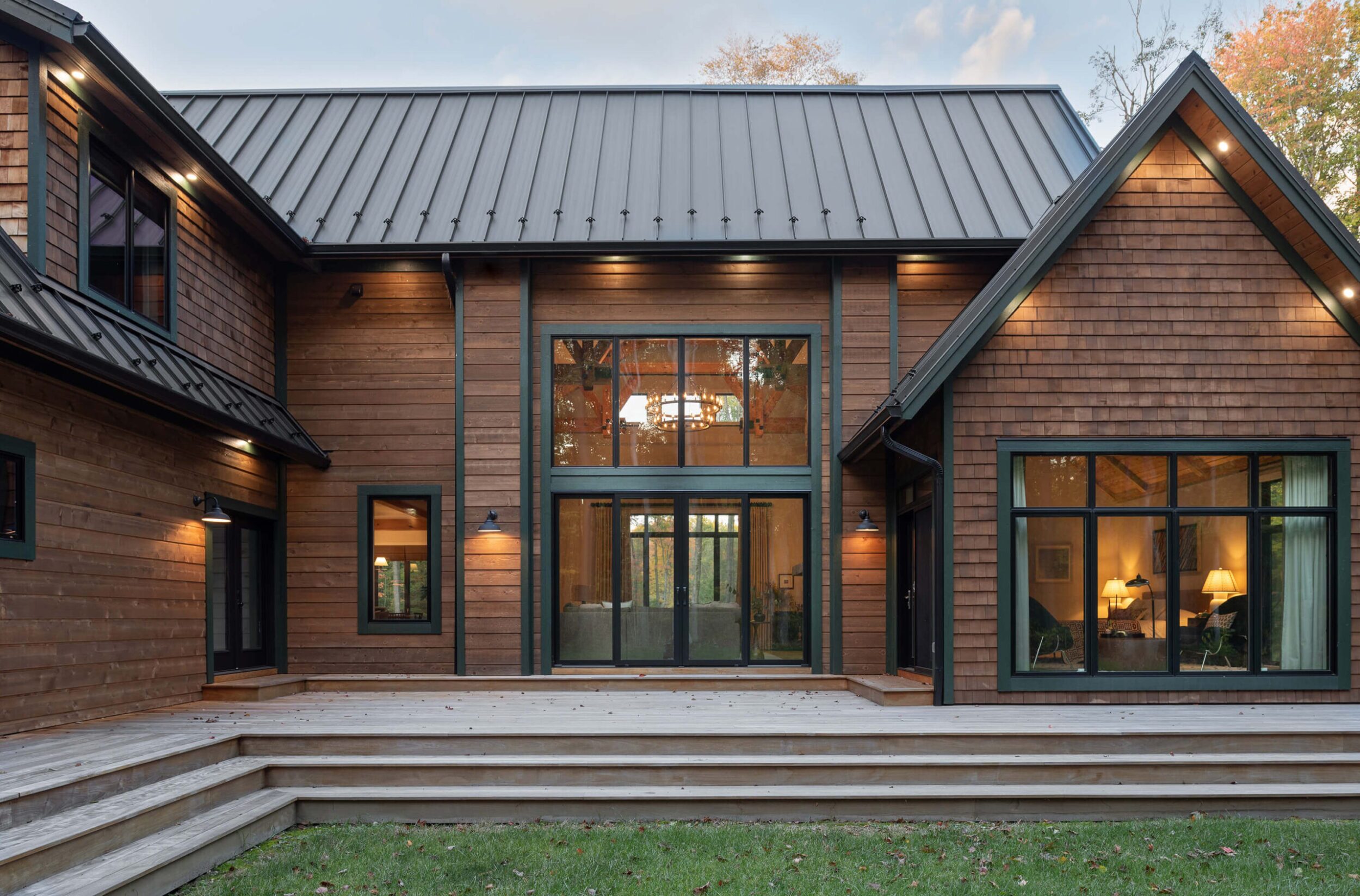Back view of a modern cabin with large windows, wooden deck, and dark roofline blending into autumn forest landscape