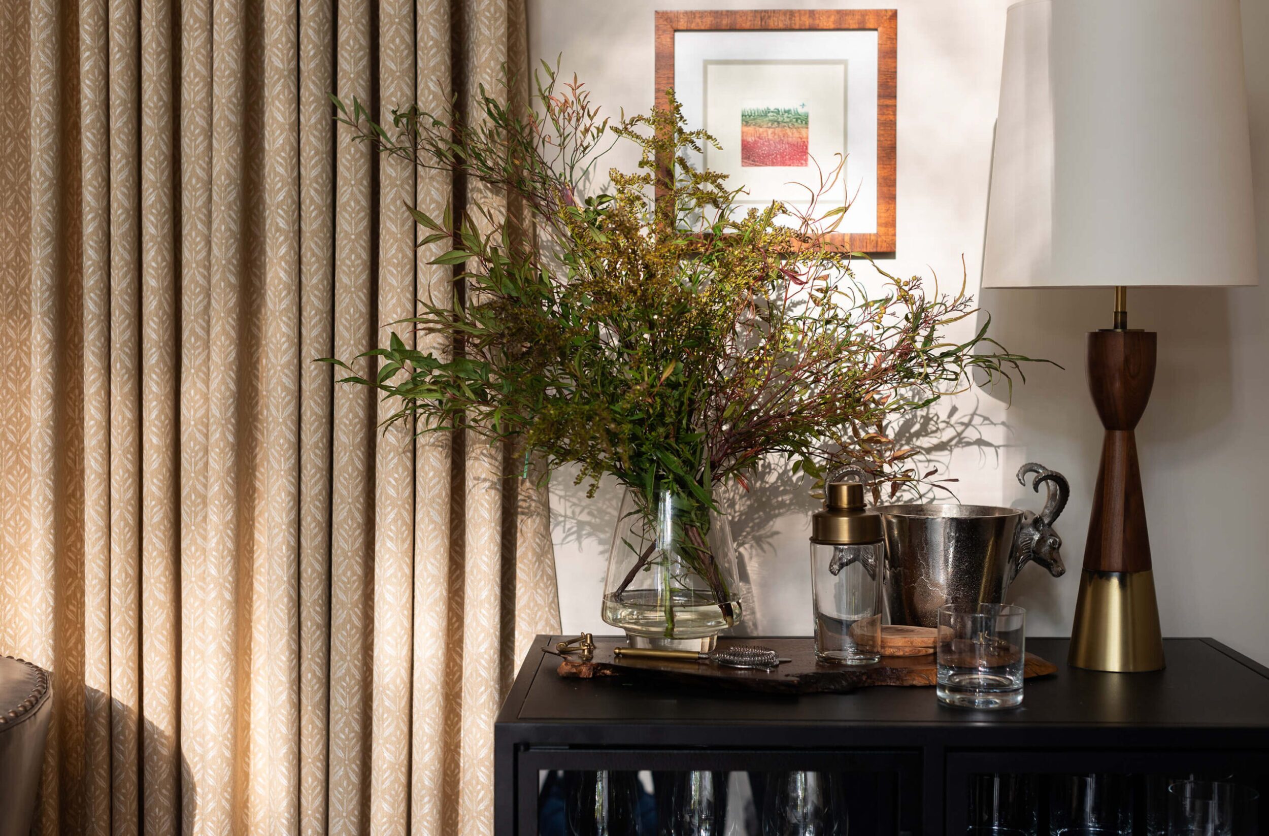 Rustic bar cabinet with greenery in a glass vase, wood lamp, and metal barware against patterned beige curtains