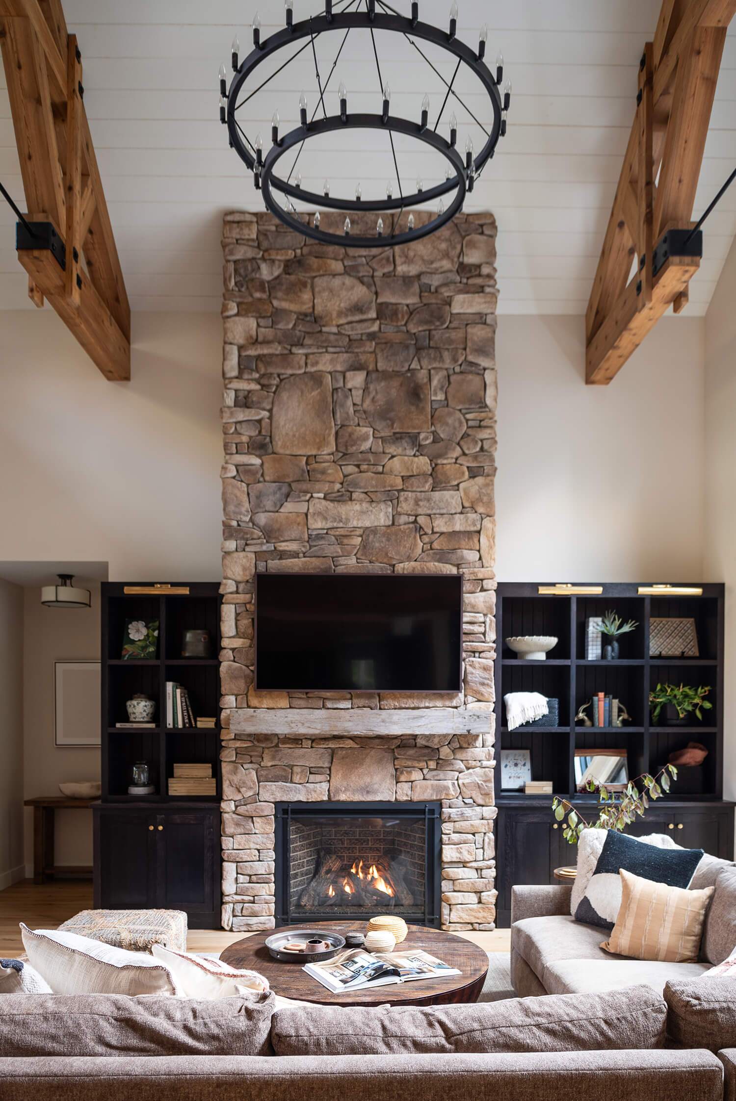 Stone fireplace with mounted TV, wooden mantel, cozy seating, dark shelves, and rustic chandelier in cabin interior