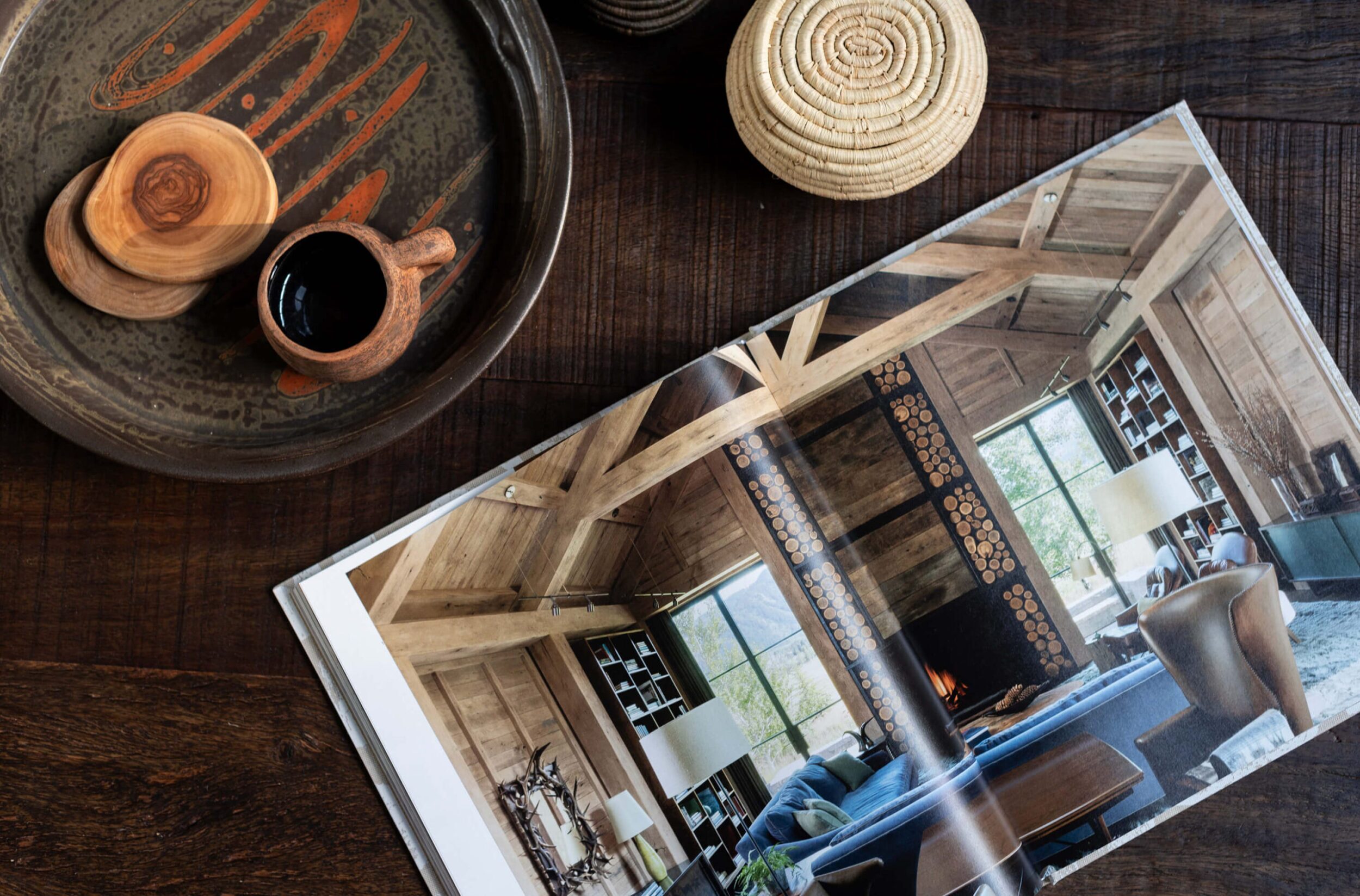 Close-up of rustic wooden table with ceramic tray, woven coaster, cup, and open magazine featuring cabin interior