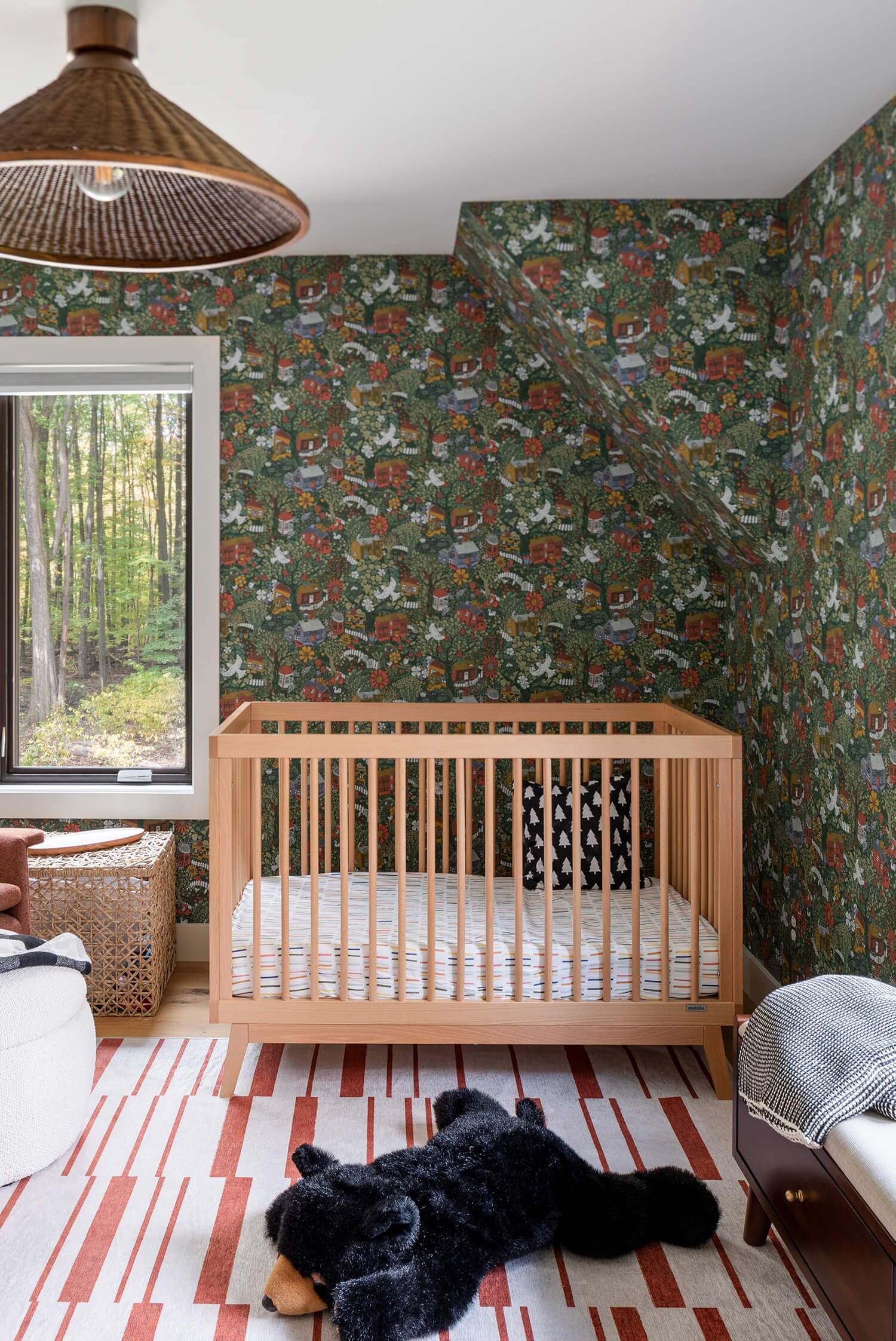 Nursery with wooden crib, forest wallpaper, red-striped rug, and large black teddy bear on floor