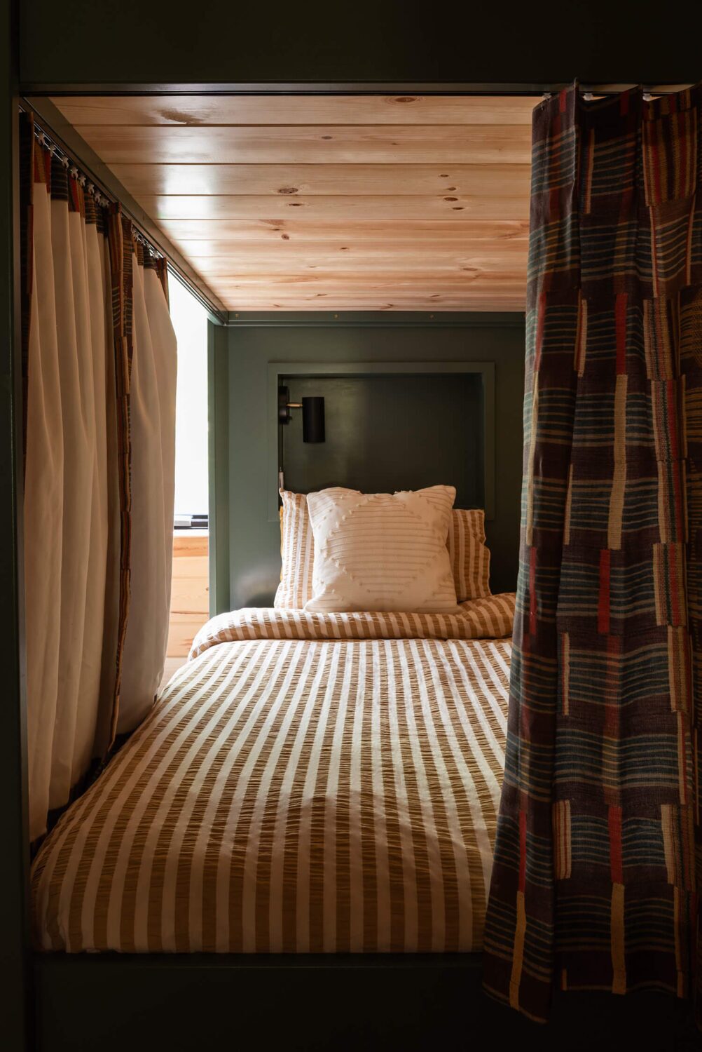 Cozy built-in bunk bed with striped bedding, accent pillow, wood ceiling, and patterned curtains in a warm modern room.